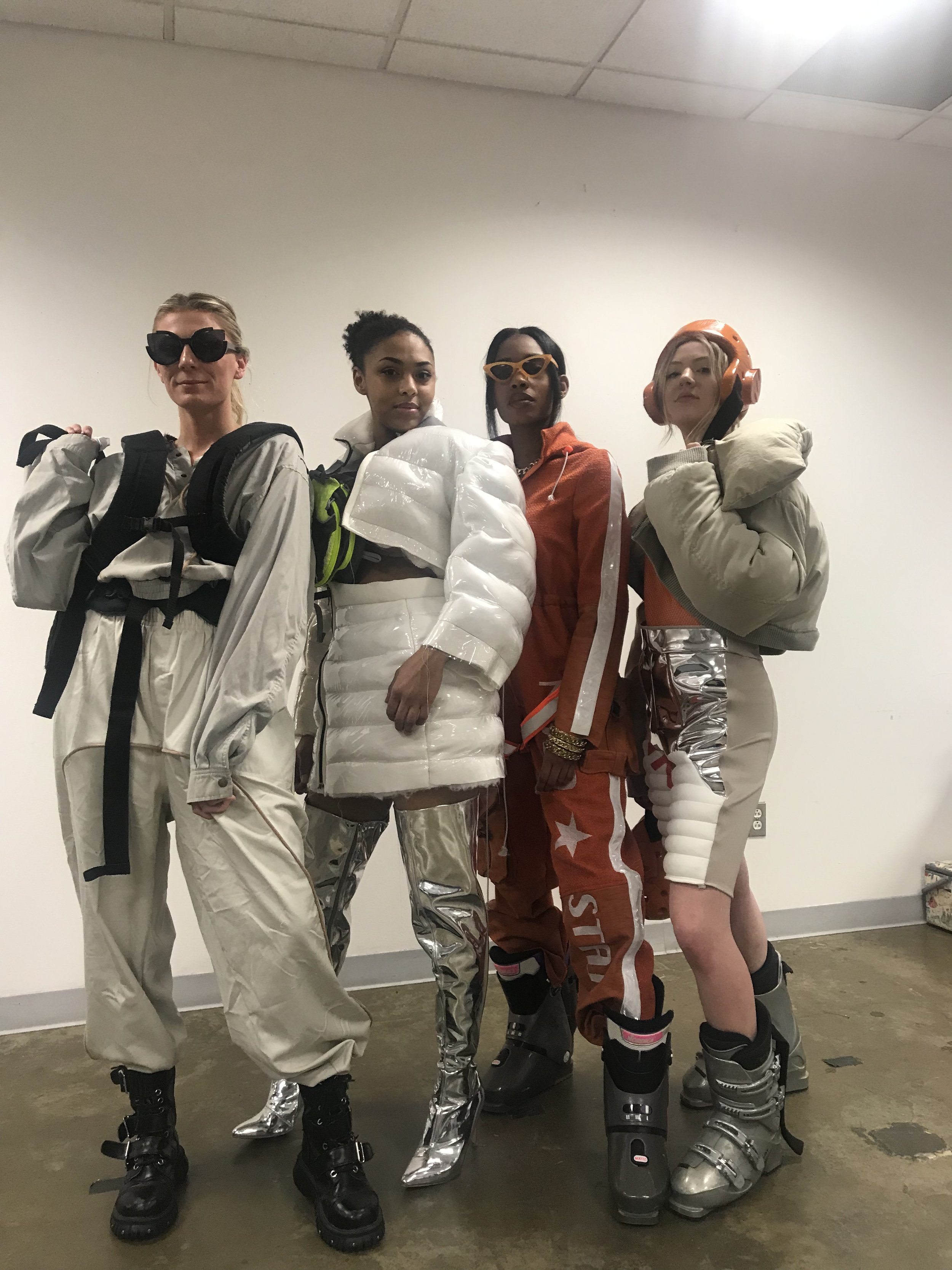 Four women wearing futuristic, space-inspired outfits and accessories posing against a plain wall.