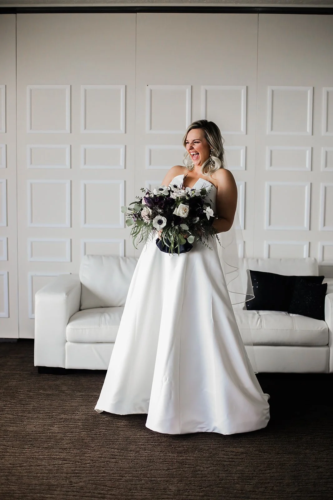 A bride wearing a white wedding gown, holding a bouquet, standing in front of a white couch and white wall, laughing enthusiastically.