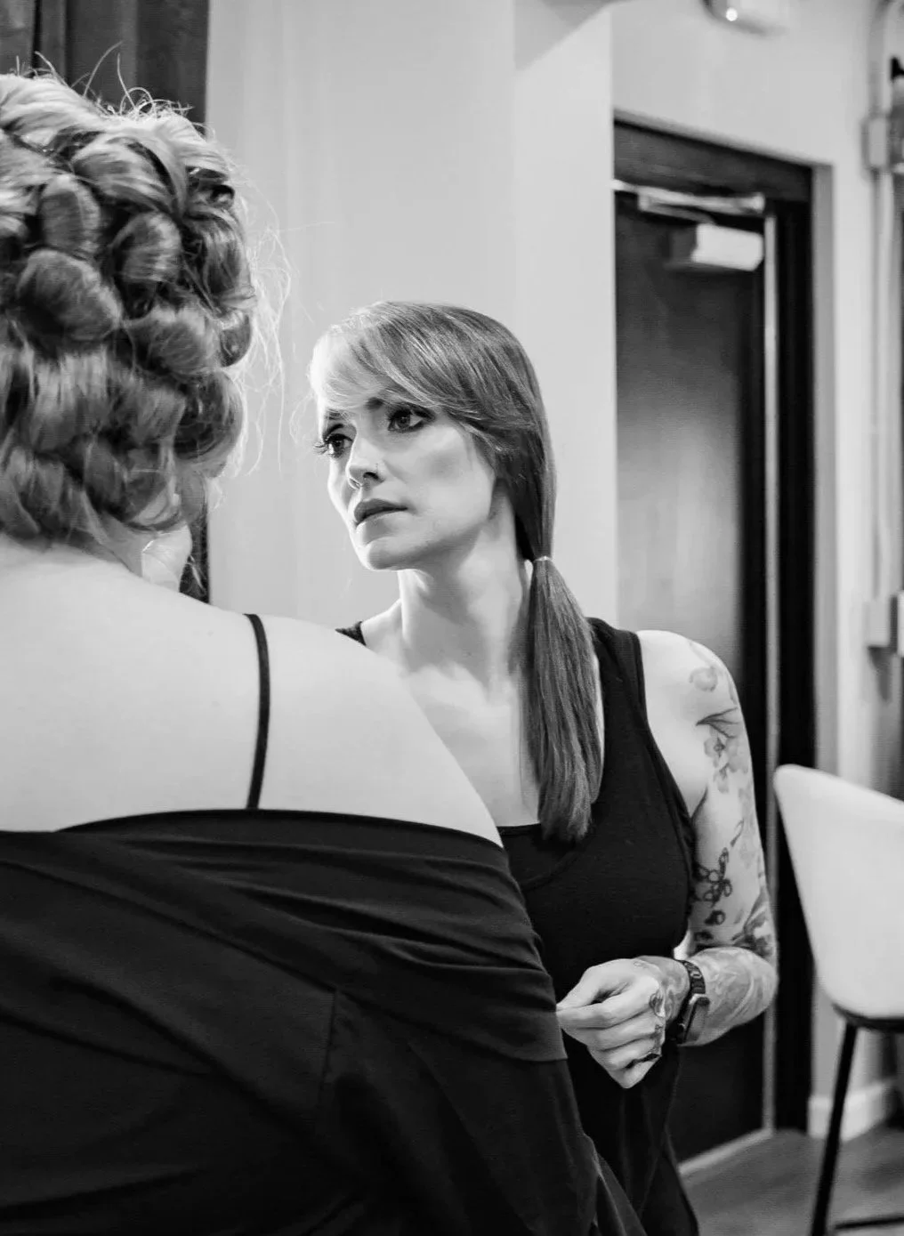 Two women, one a bride with her back to the camera and her hair set in curls, and the other a makeup artist with straight hair and tattoos doing her makeup.
