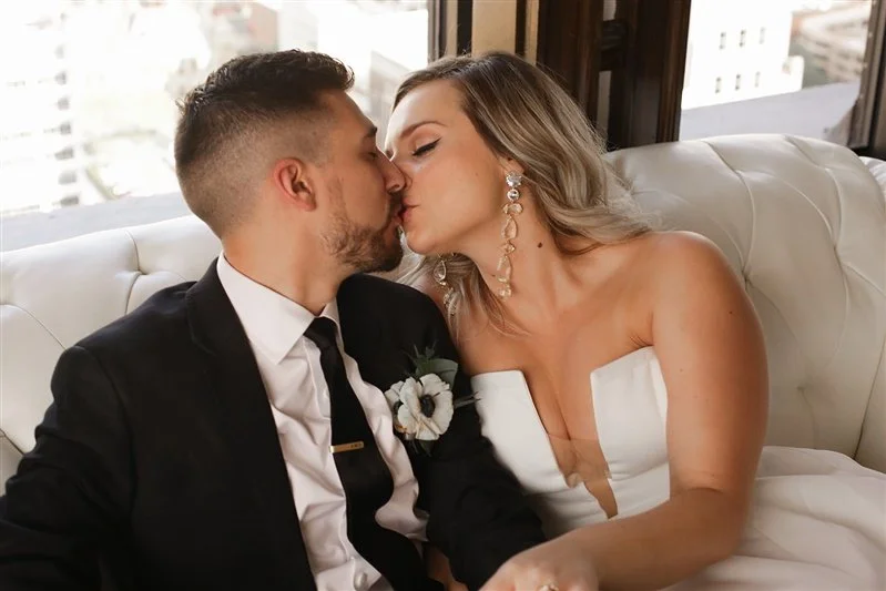 A groom wearing a black and white suit and a bride wearing a white wedding gown sitting on a white tufted couch, holding hands, kissing.