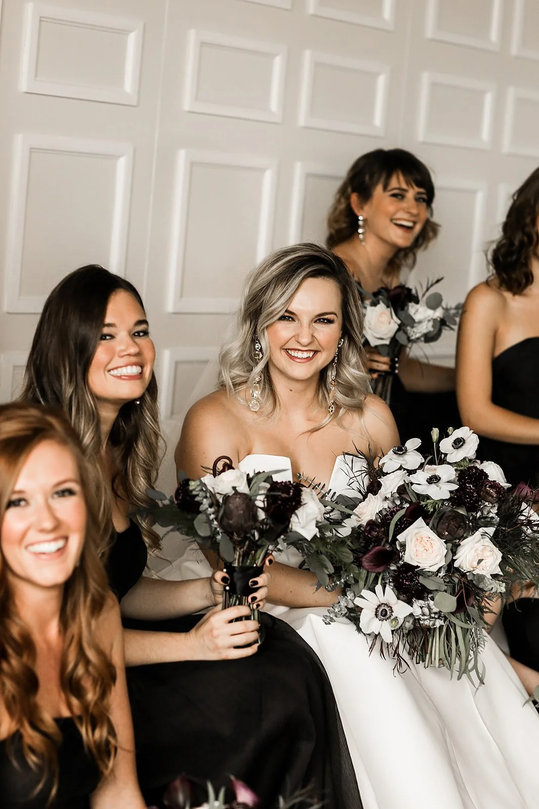 A bride wearing a white wedding gown with all her bridesmaids wearing black dresses. Everyone is holding bouquets and sitting in front of a white wall.