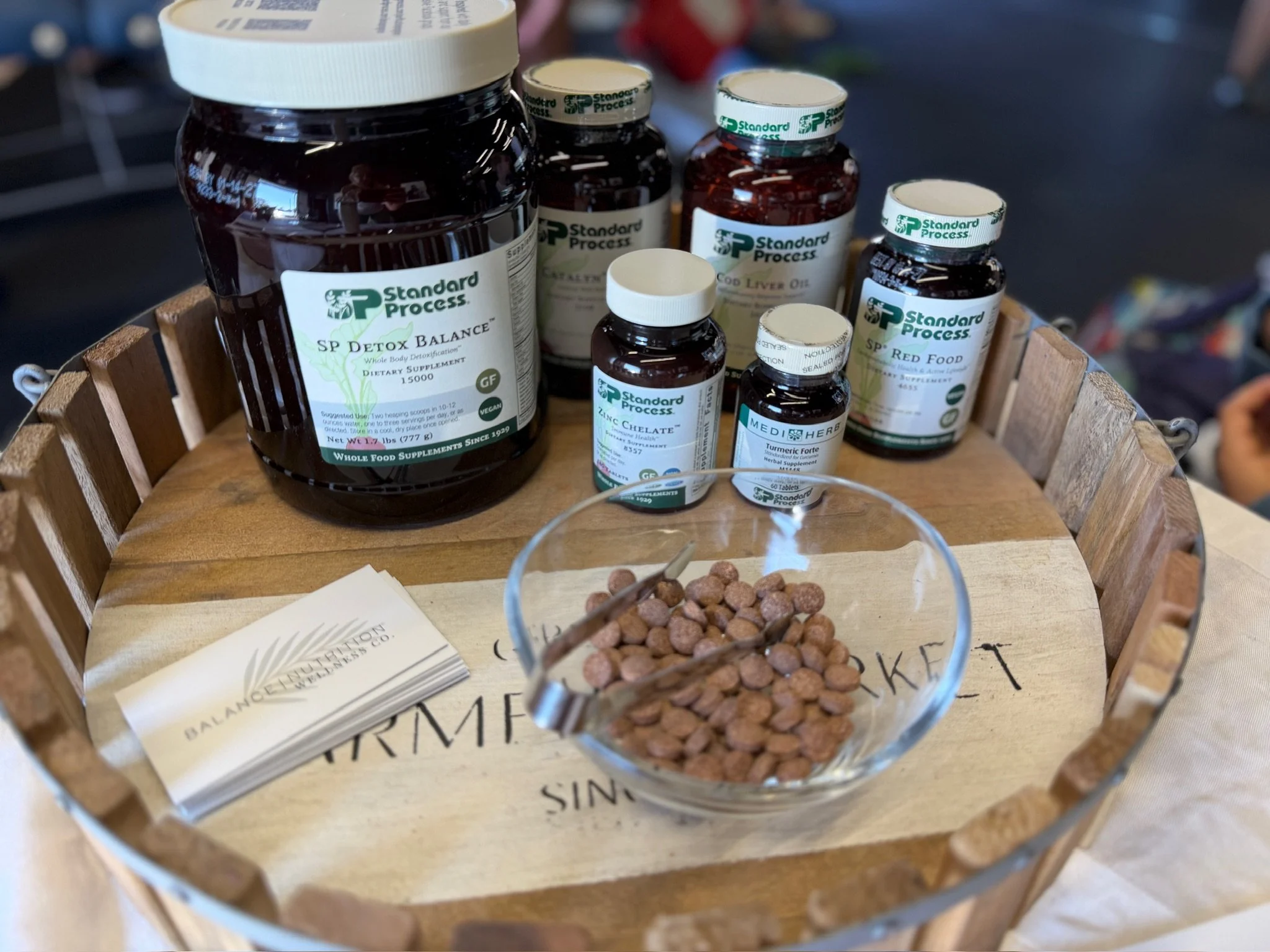Display of dietary supplement bottles arranged in a wooden basket, including products labeled 'SP Detox Balance,' 'SP Liver Oil,' 'SP Zinc Chelate,' 'SP Turmeric Forte,' and 'SP Red Food,' along with a glass bowl filled with small, round, brown tablets, and some informational pamphlets.