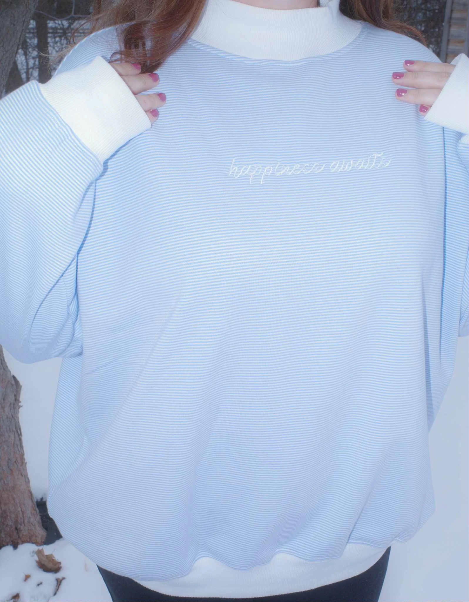 GIRL WEARING BLUE AND WHITE STRIPED MOCKNECK WITH WHITE EMBROIDERED TEXT THAT READS 'HAPPINESS AWAITS'