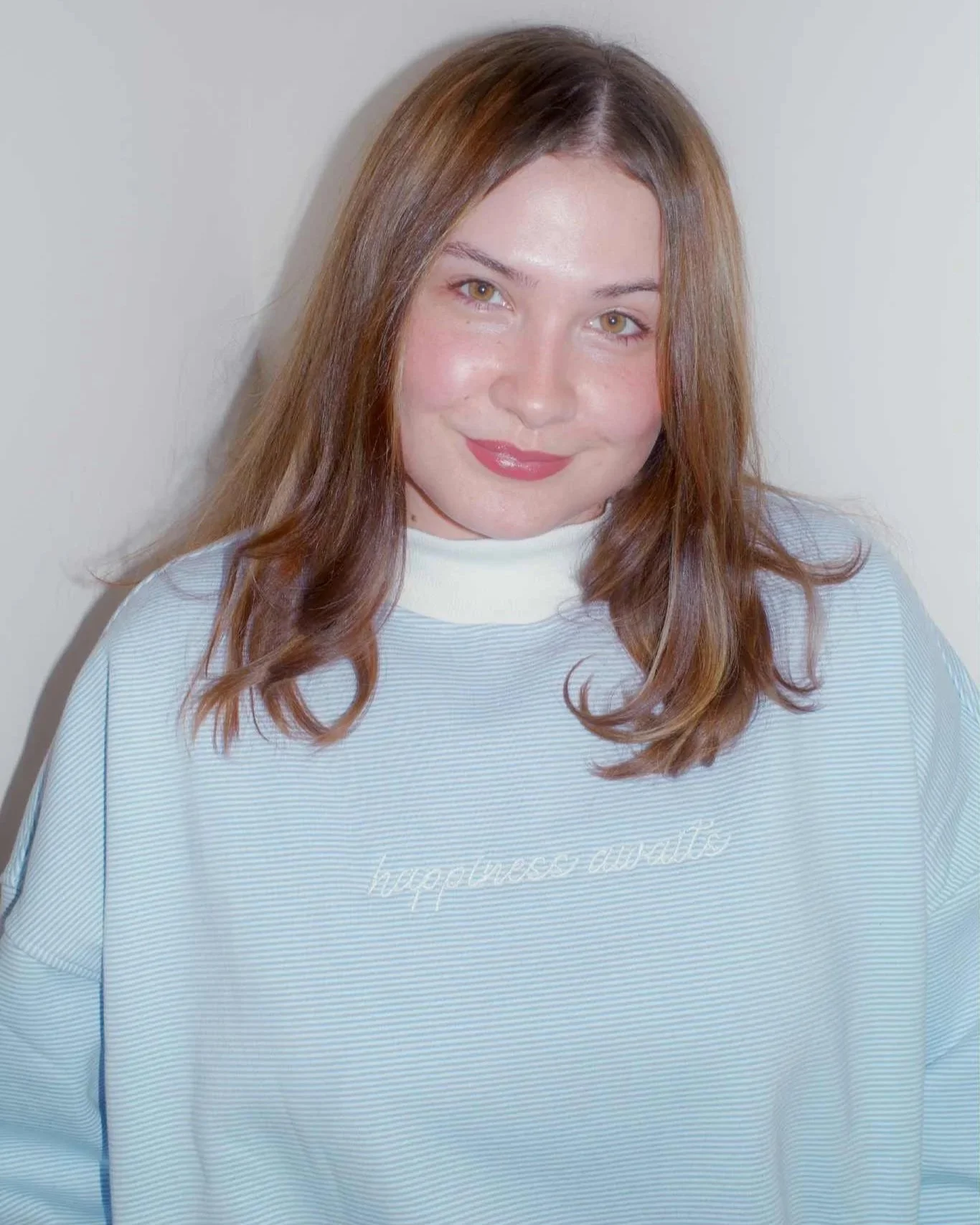 GIRL WEARING BLUE AND WHITE STRIPED MOCKNECK WITH WHITE EMBROIDERED TEXT THAT READS 'HAPPINESS AWAITS'