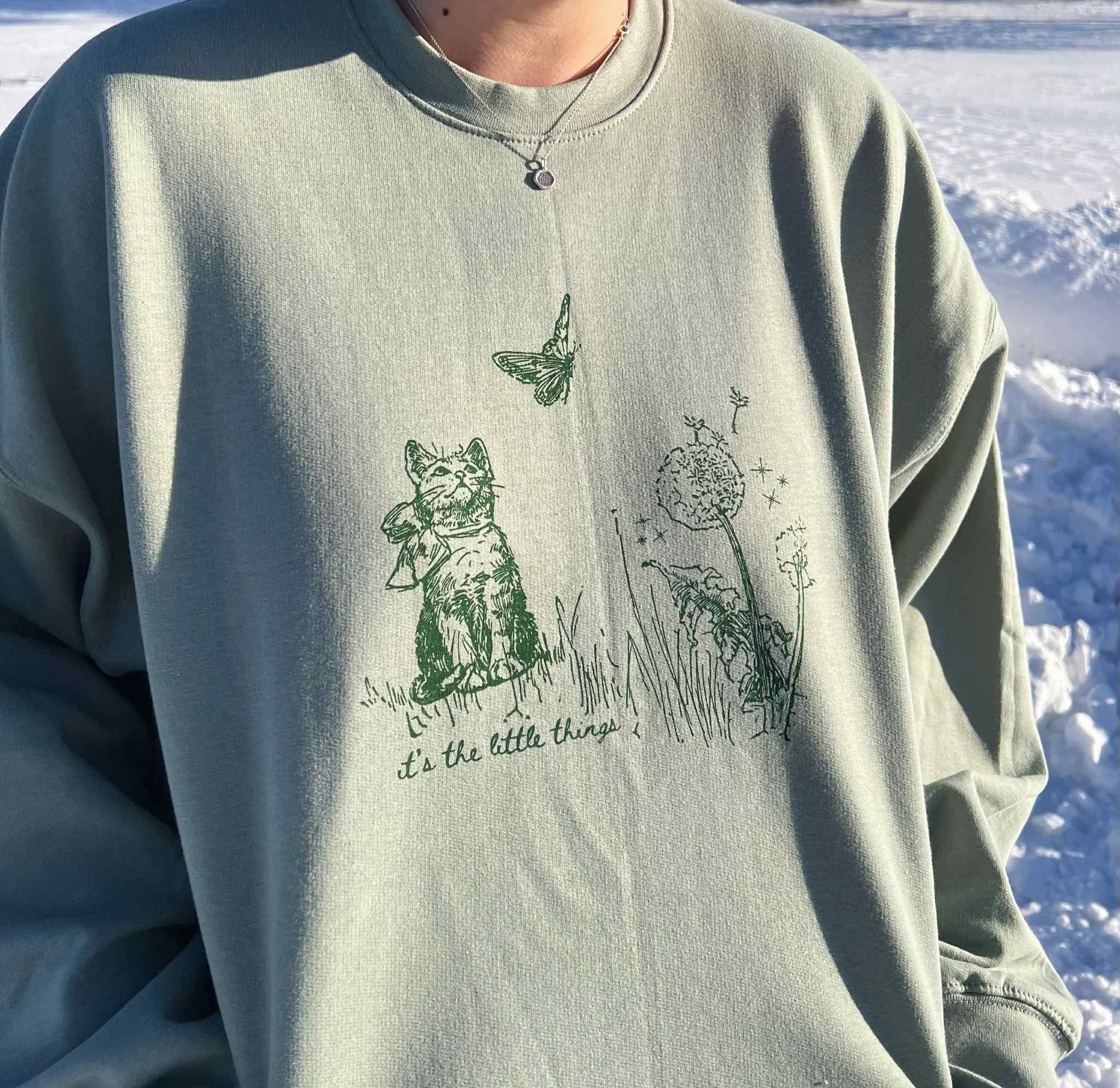 SAGE GREEN SWEATSHIRT WITH ILLUSTRATION OF CAT AND BUTTERFLY WITH TEXT THAT READS 'ITS THE LITTLE THINGS' BELOW