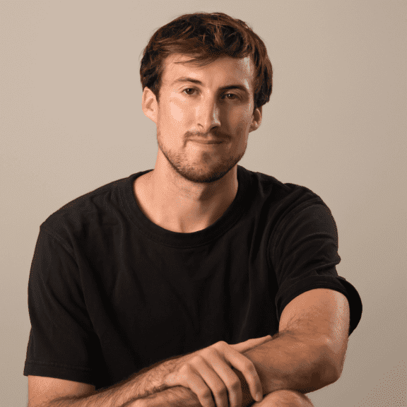 A young man with blonde hair, blue eyes, and a beard, smiling, wearing earrings and a necklace, against a dark blue background.