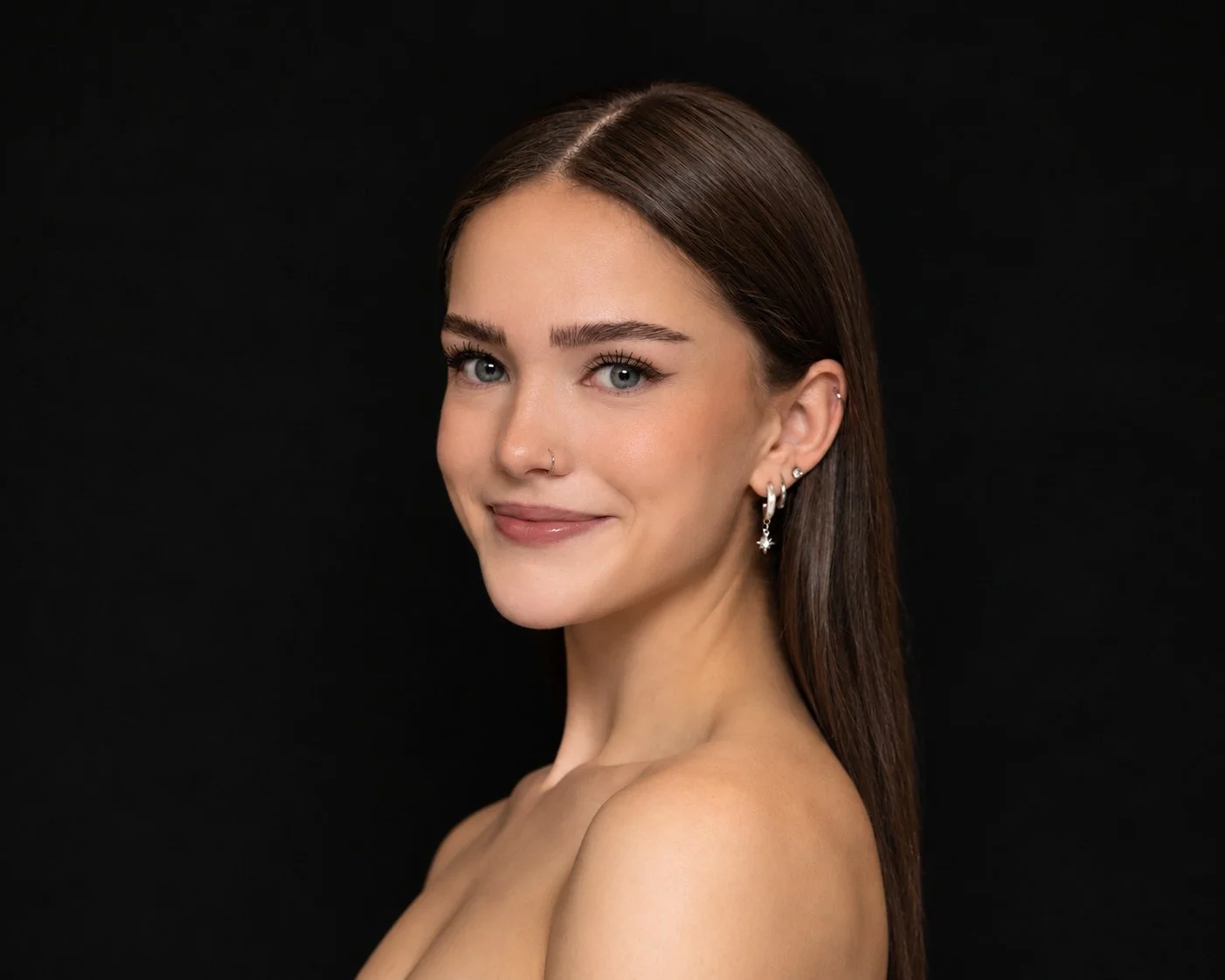 A woman with fair skin, dark brown hair styled in a sleek, knowing bun, blue eyes, and well-defined eyebrows. She is wearing subtle makeup, including winged eyeliner and light pink lipstick, and has multiple earrings in her left ear, including a hoop and several studs. She is posed against a plain white background with an elegant, confident expression.