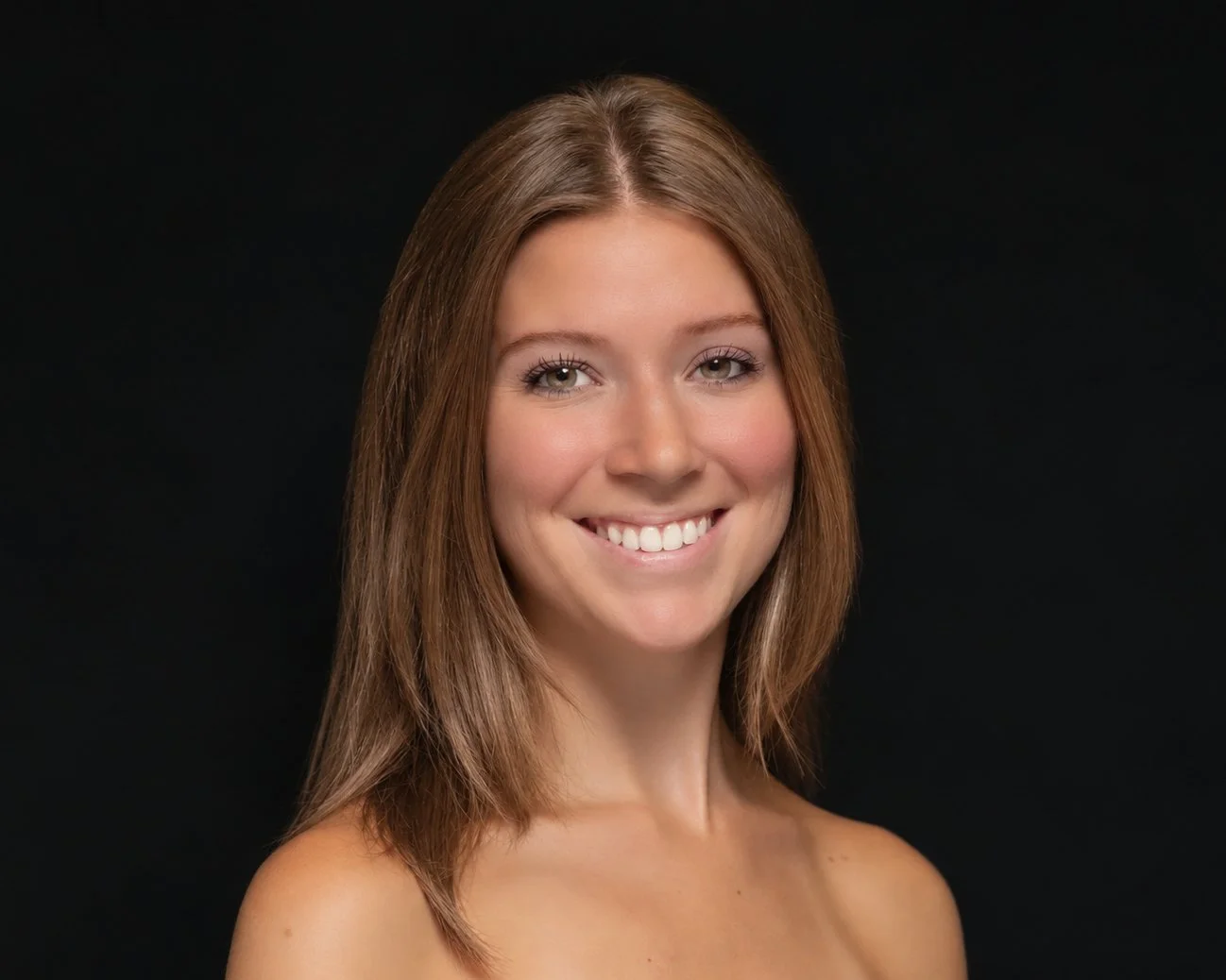 A young woman with long brown hair, light skin, and green eyes smiling at the camera, wearing gold earrings and a sleeveless top.
