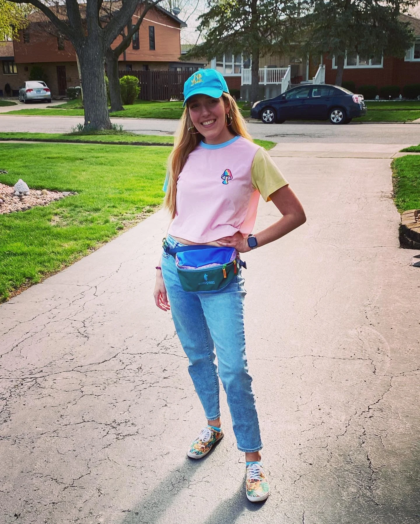 A young woman with long blonde hair standing on a cracked asphalt driveway in a residential neighborhood. She is smiling, wearing a colorful pastel t-shirt, blue jeans, floral sneakers, a blue cap, and a waist pack. There are green lawns, trees, and houses in the background.