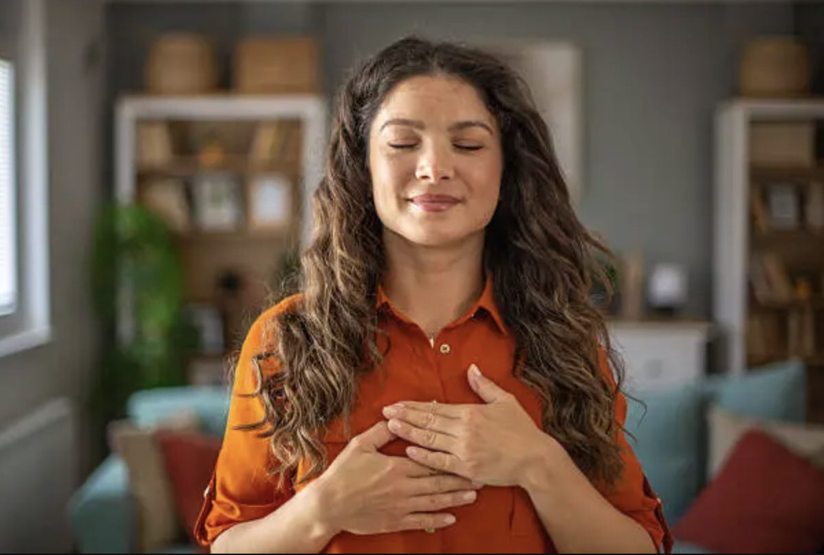 Woman with long curly hair wearing an orange shirt, eyes closed, smiling, with her hands over her chest.
