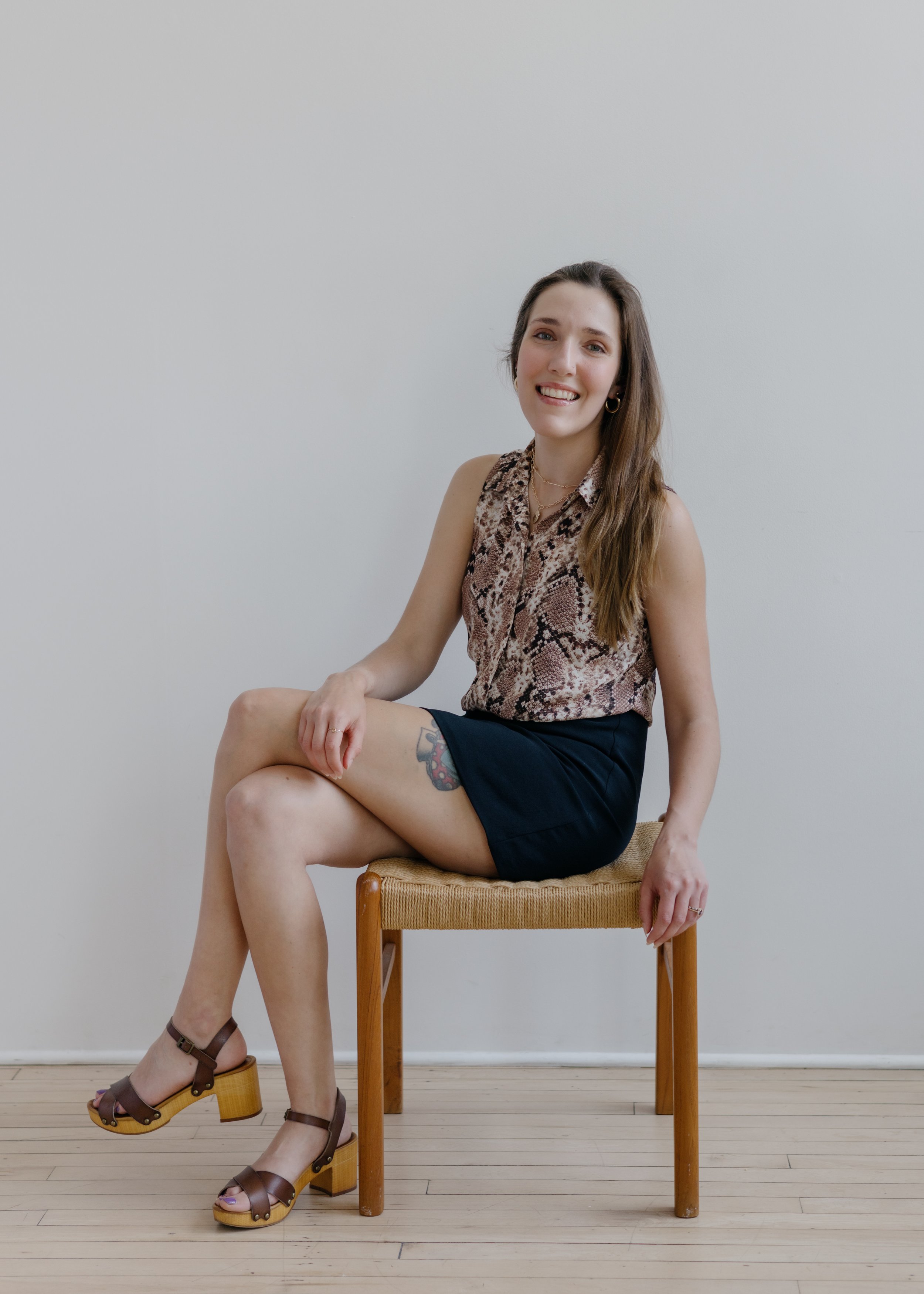 A woman with long brown hair smiling, sitting on a wooden stool with a woven seat against a plain white wall. She is wearing a snake print sleeveless blouse, dark shorts, and brown platform sandals with wooden heels, showing a tattoo on her thigh.