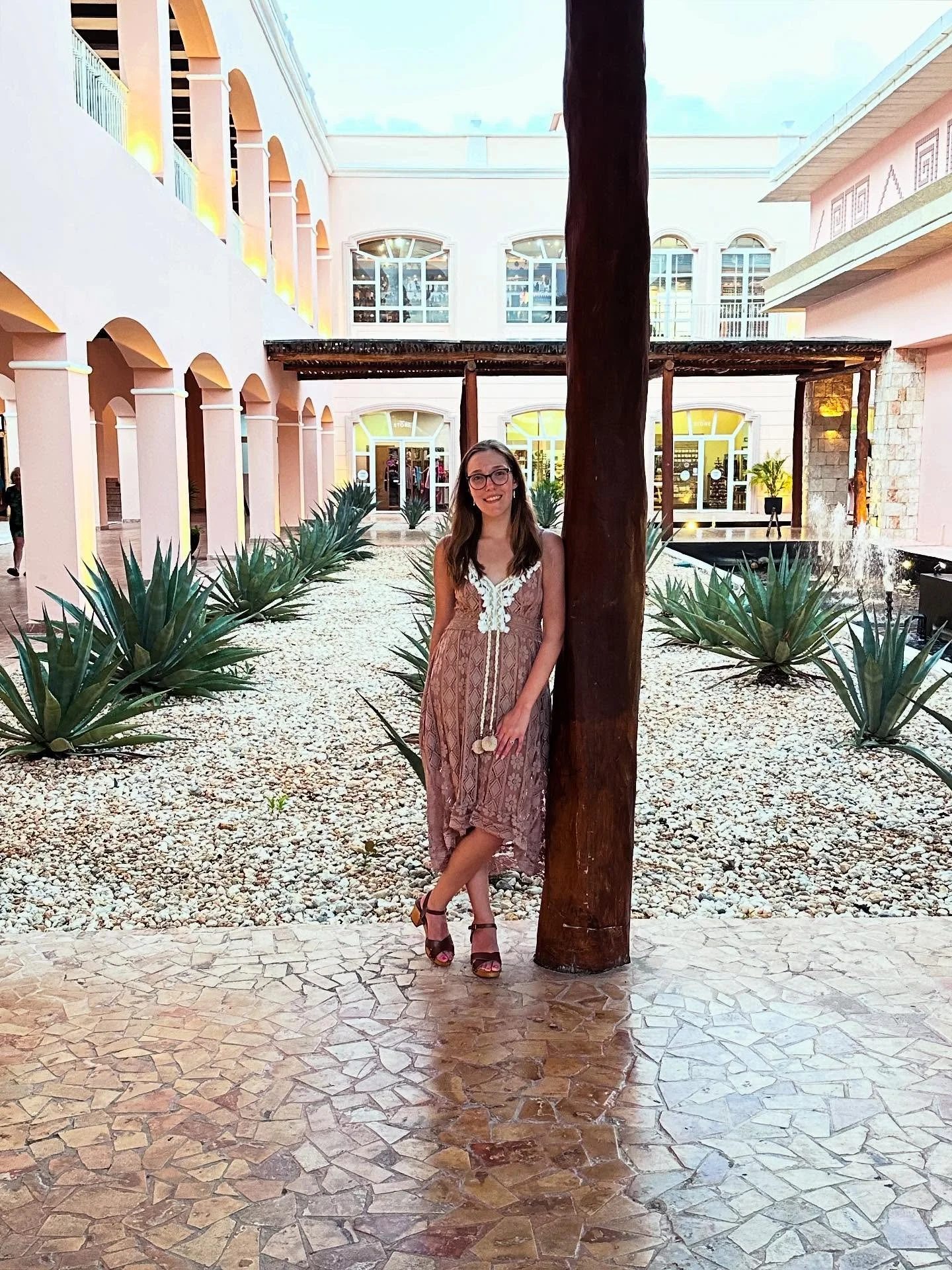 A woman stands with her back against a tree in a courtyard with pink and white buildings, agave plants, and a fountain.