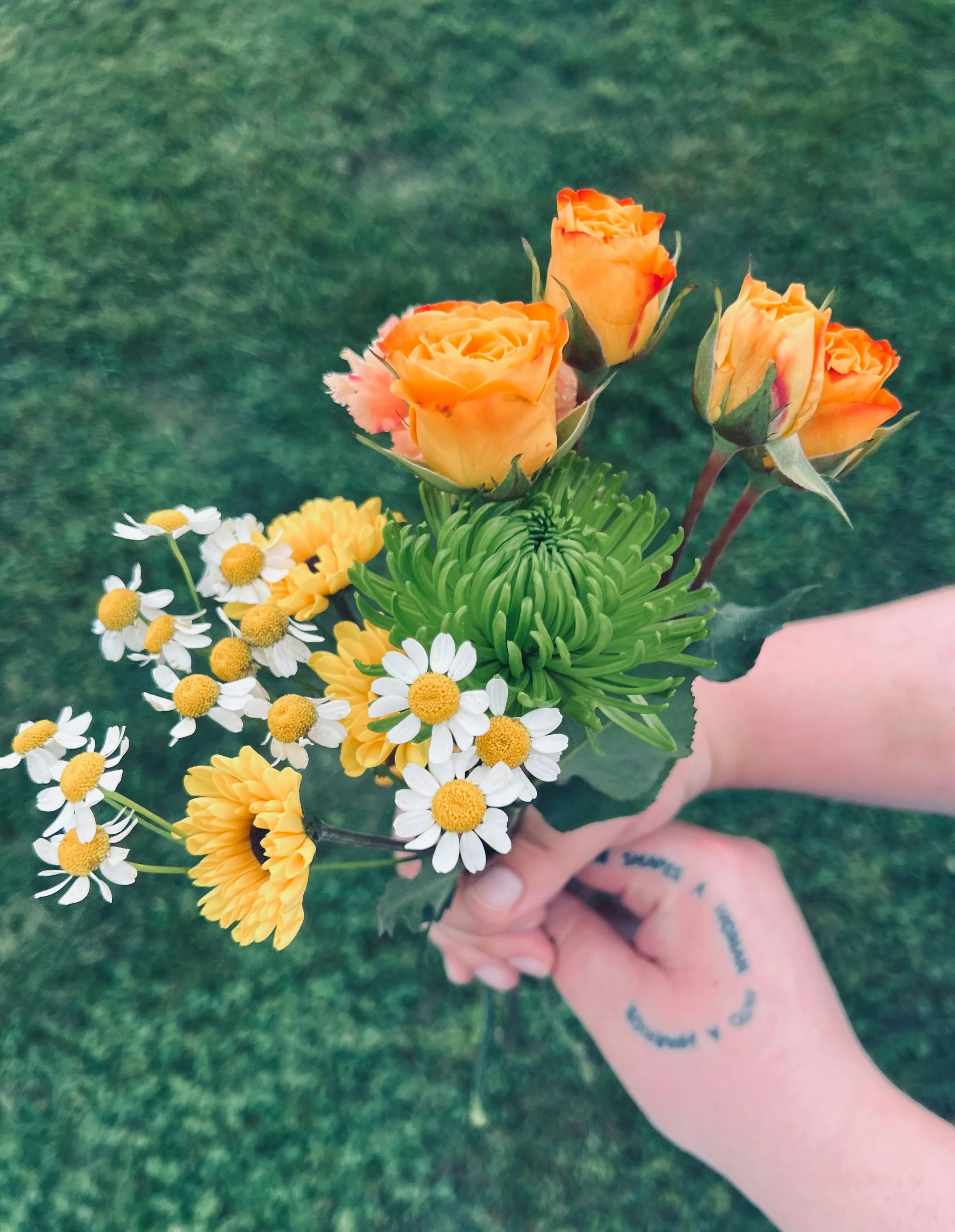 A person holding a bouquet of orange roses, white daisies, and yellow flowers against a grassy background.