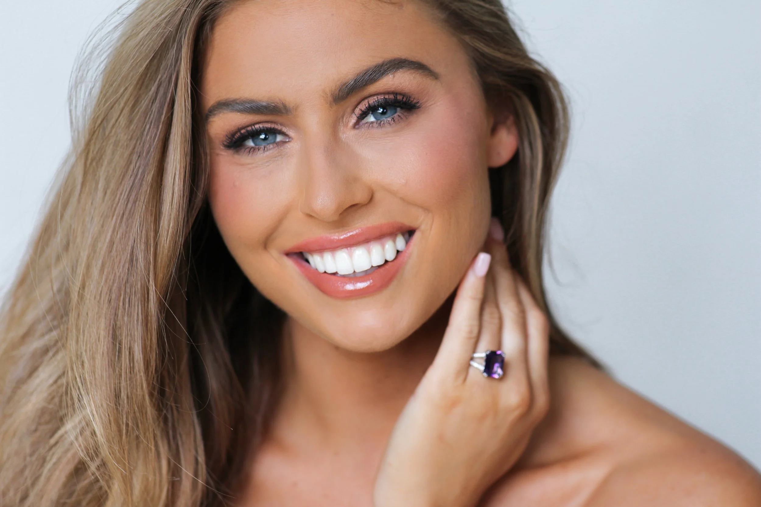 Miss Arizona USA  with her Helmich Luxury Jewels ring. Close-up of a smiling woman with long, wavy blonde hair, blue eyes, and makeup. She is touching her face with her left hand, wearing a ring with a purple gemstone.