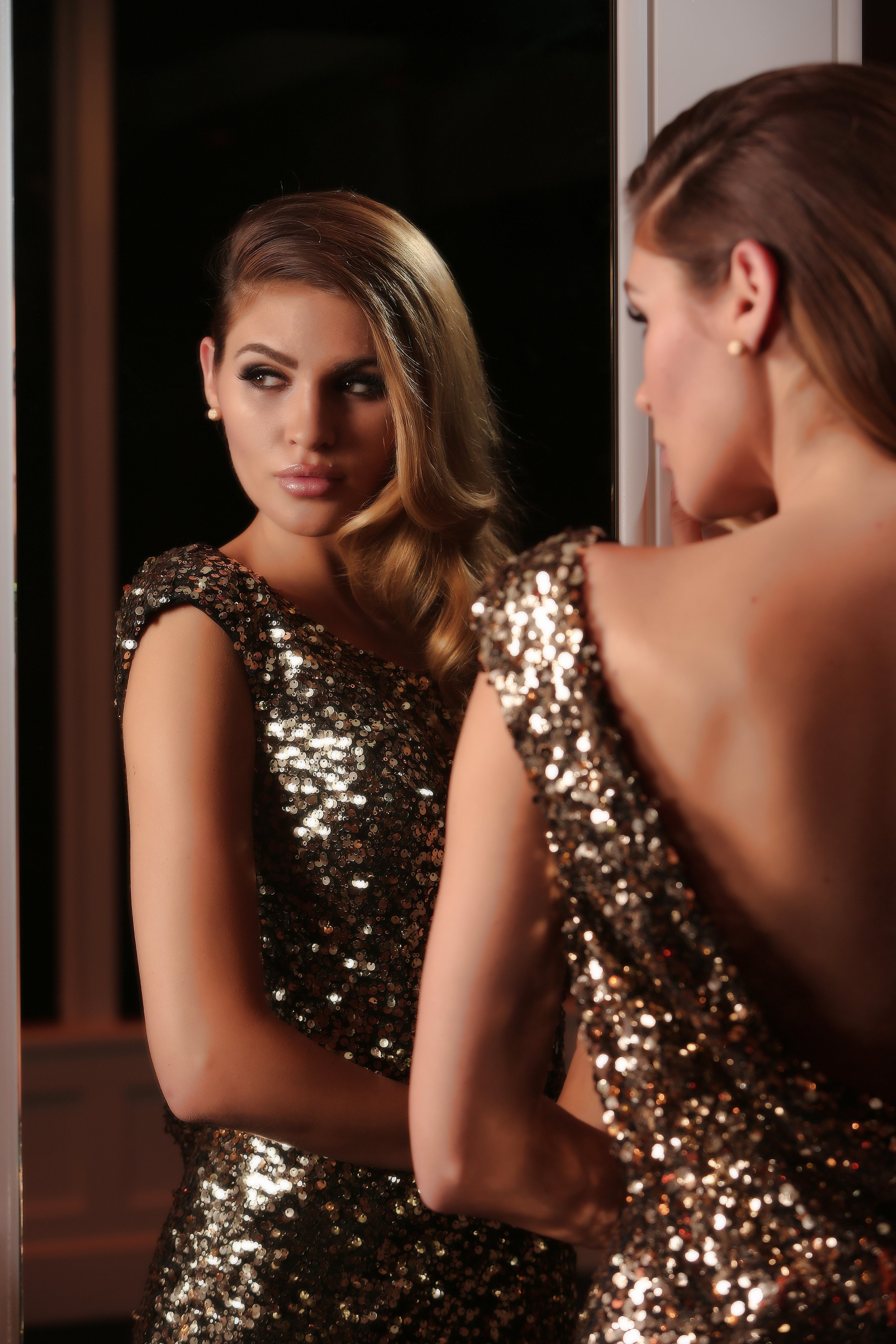 A woman with long brown hair in a side braid wearing a sleeveless, sequined black dress looking at her reflection in a mirror.