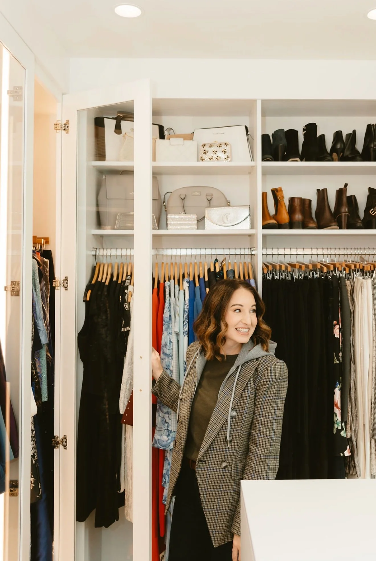 A woman smiling in a dressing room among clothing racks and shelves with shoes and accessories.