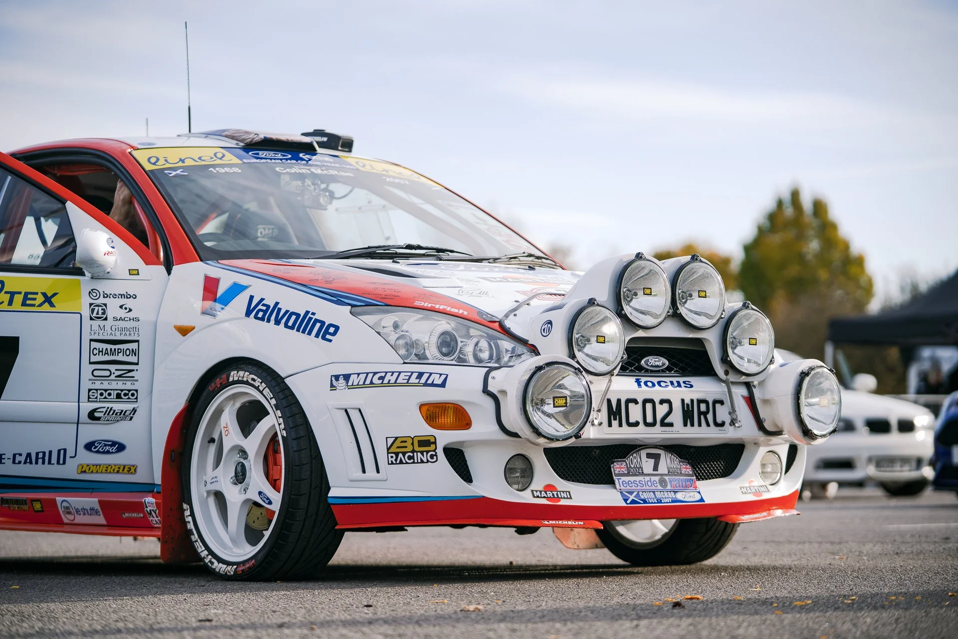 Race car with multiple headlights and sponsor stickers, parked on a paved surface with trees and tents in the background.