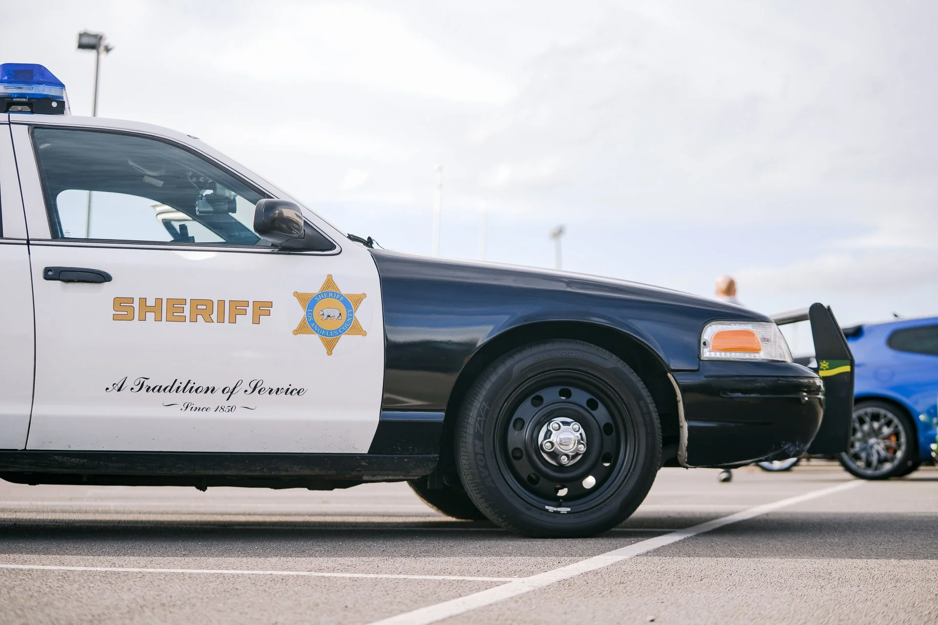 Los Angeles County sheriff's patrol car parked in a lot with a blue car in the background under a cloudy sky.