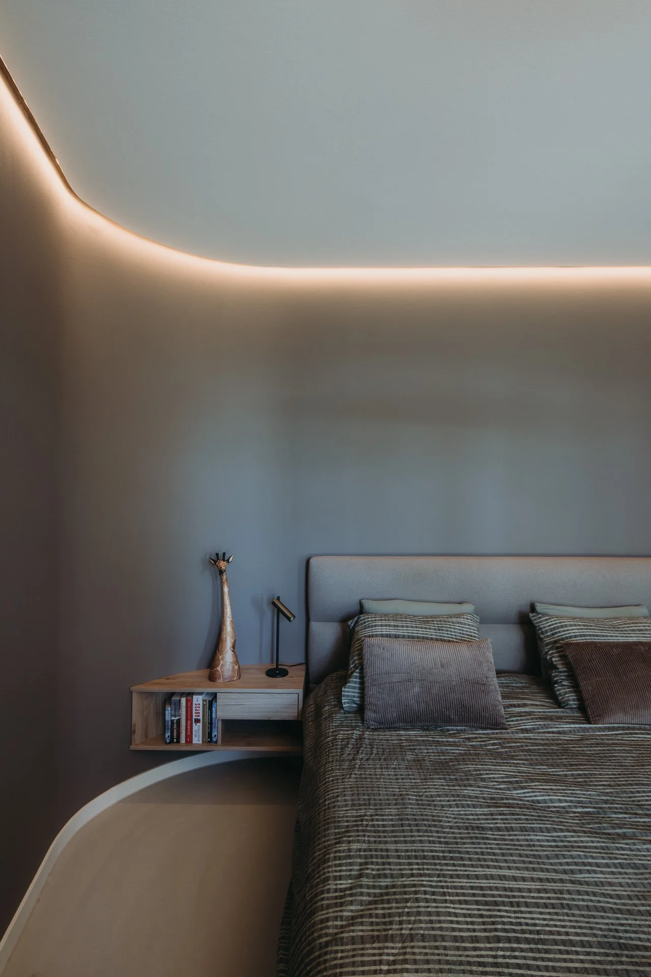 Warme taupe kleurige gebogen wand achter bed, met strijklicht van lichtlijn in plafond.