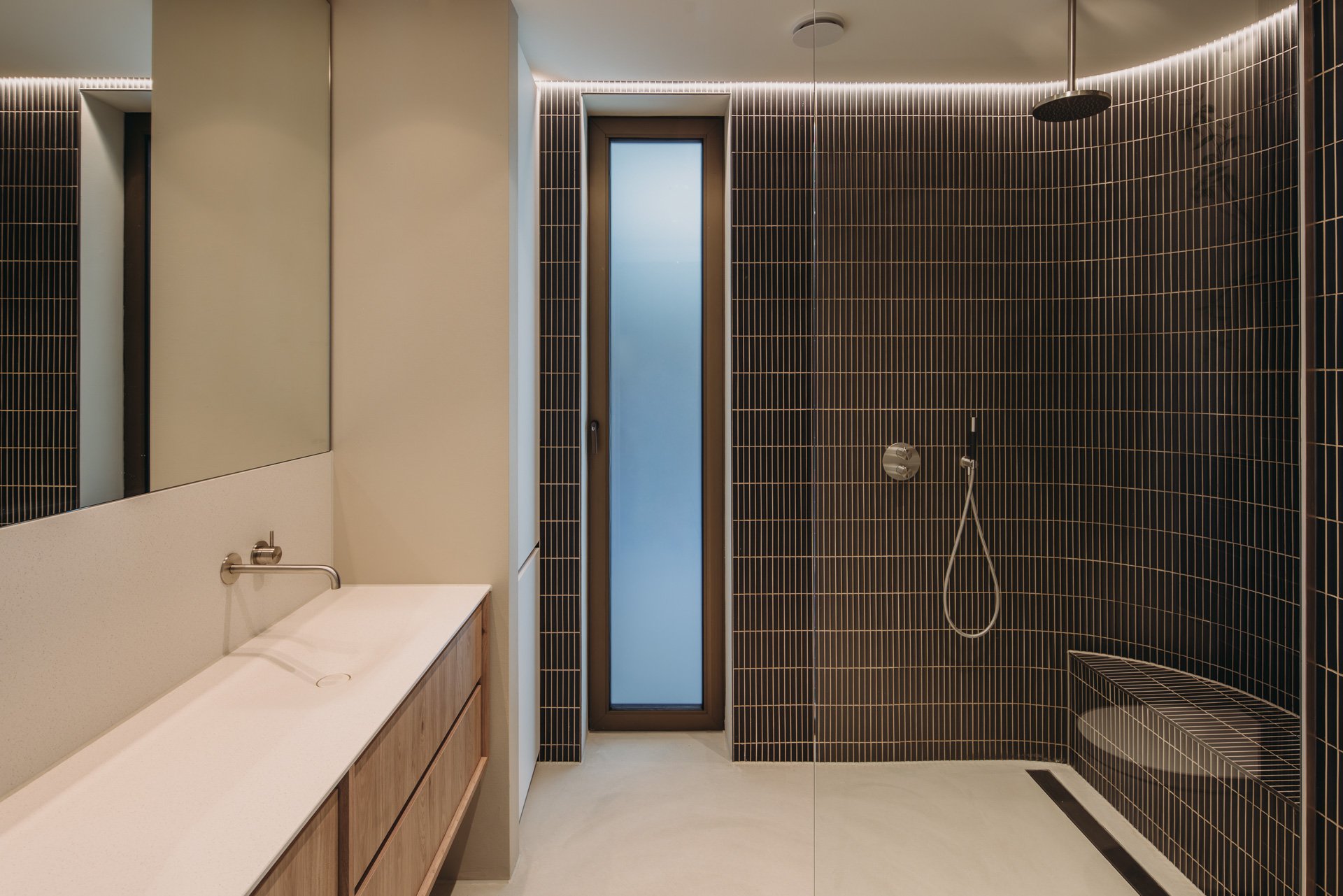 Luxe badkamer met warme donkerbruine betegelde wanden en moderne minimalistisch ontworpen houten wastafelmeubel tegen contrasterend licht beige wand.