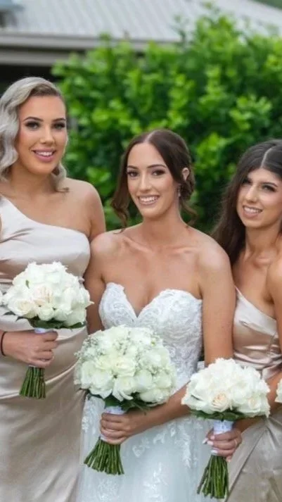 Three women in wedding and bridesmaid dresses smiling and holding bouquets of white flowers outdoors with green trees in the background.