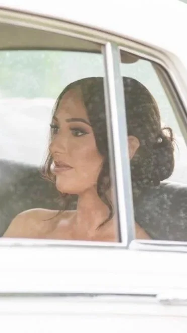 A woman with dark, curly hair and makeup sitting inside a car, seen through the window.