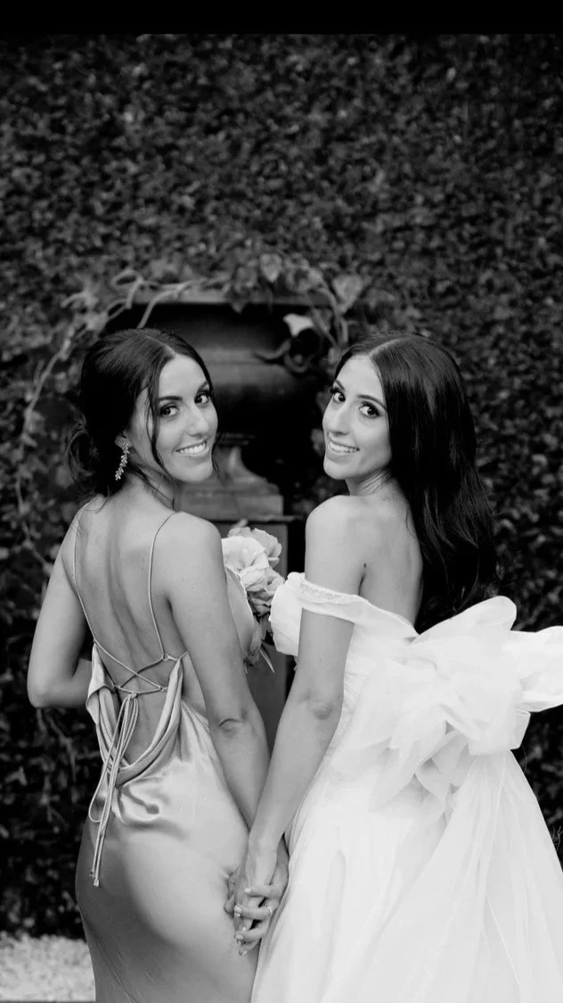 Two women dressed in formal gowns holding hands and smiling, with a hedge and decorative iron gate in the background, in black and white.
