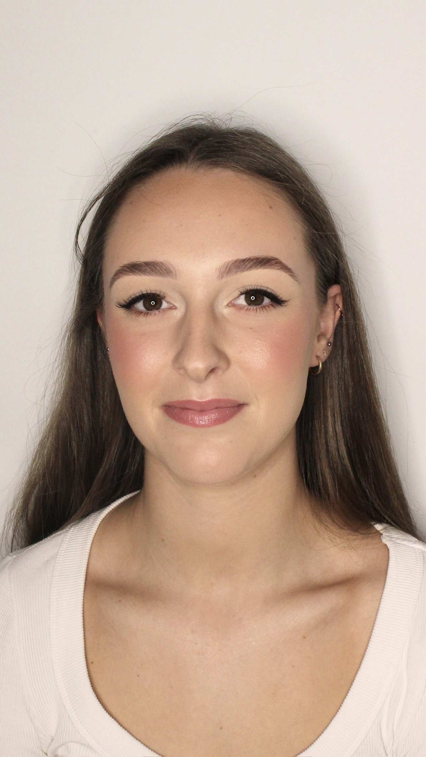 Close-up portrait of a young woman with long brown hair, wearing makeup, earrings, and a white top, standing against a plain white background.
