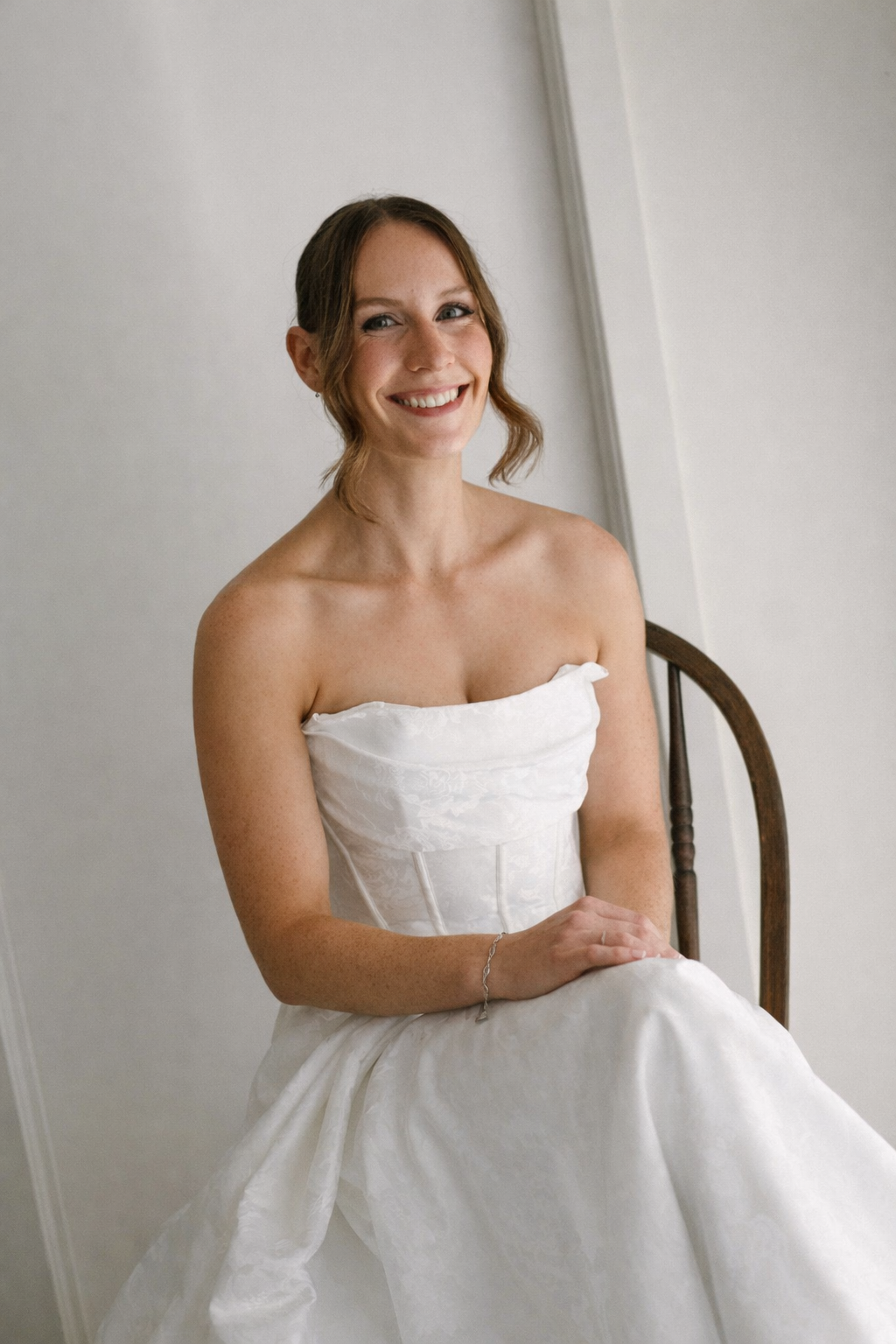A smiling woman in a white dress sitting on a wooden chair.