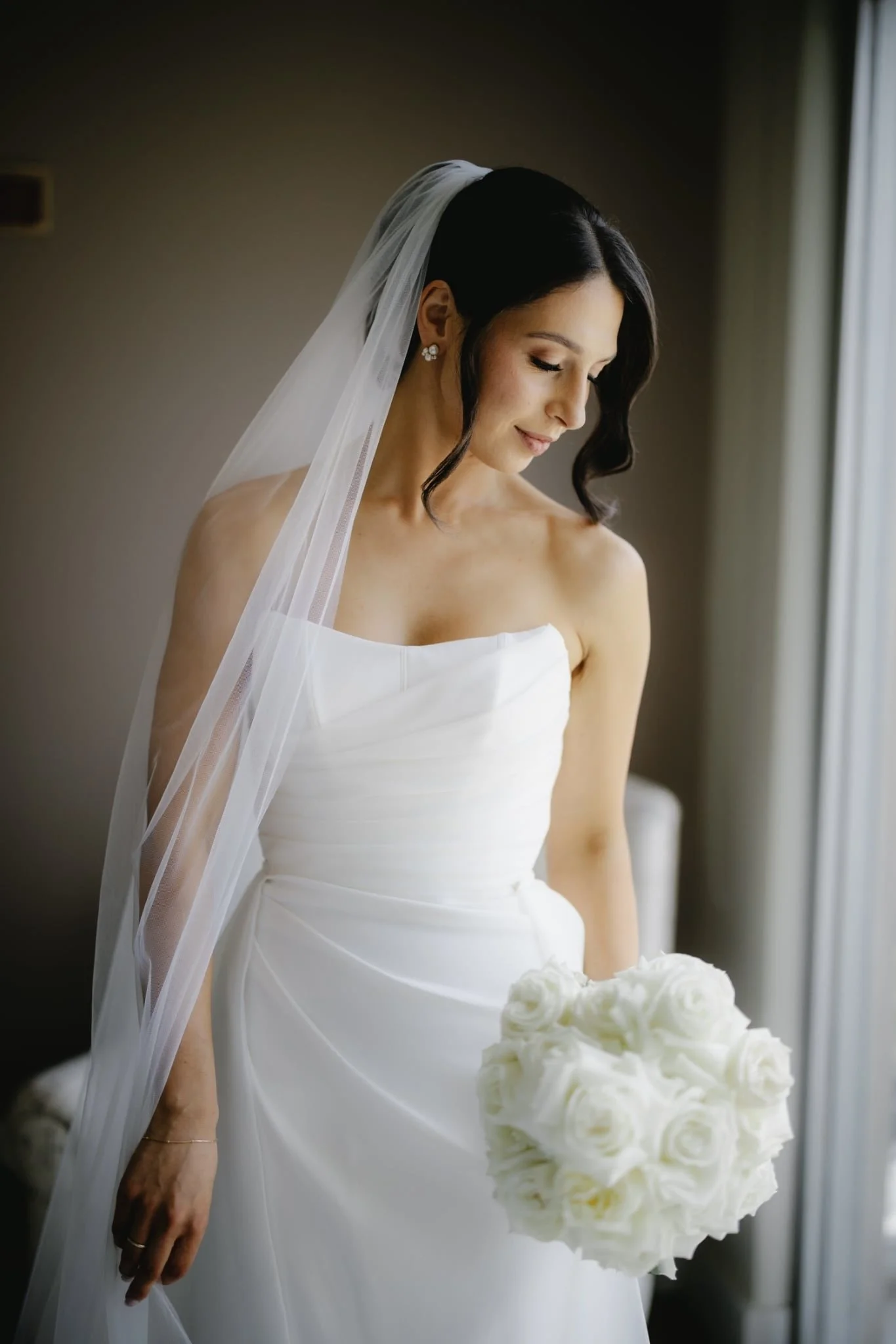 A bride in a white wedding dress holding a bouquet of white roses, standing near a window with natural light.