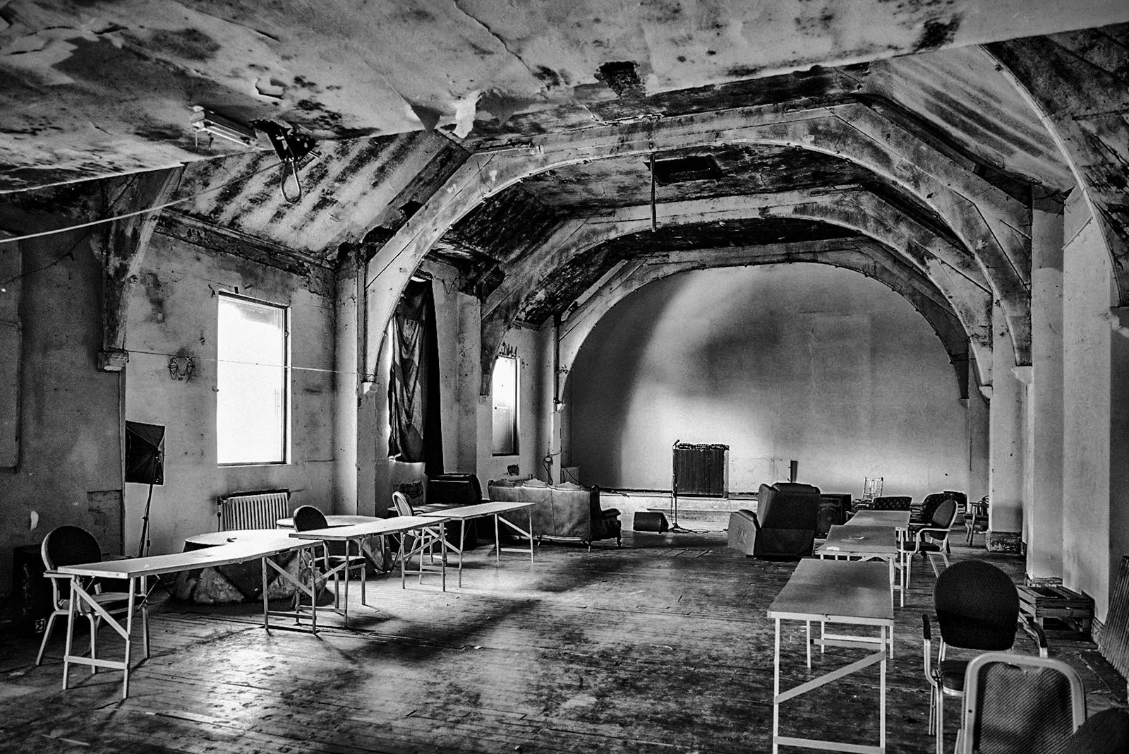 An abandoned, possibly historic, large indoor space with arched ceiling, worn walls, and scattered furniture including tables, chairs, and a couch. The space has two windows letting in light.