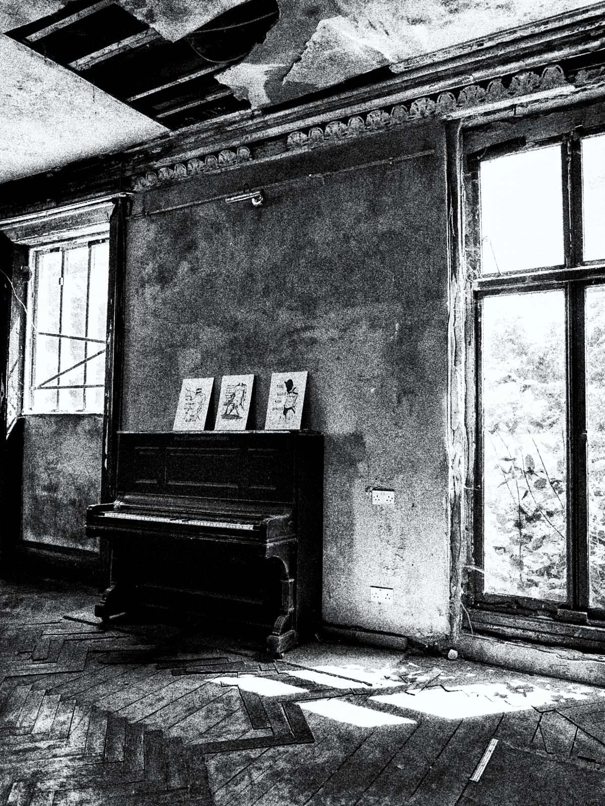 An old, abandoned room with a piano and three pictures on top of it. The room has peeling paint, weathered walls, and cracked ceiling with missing parts. There are two large windows allowing sunlight to cast shadows on the floor.