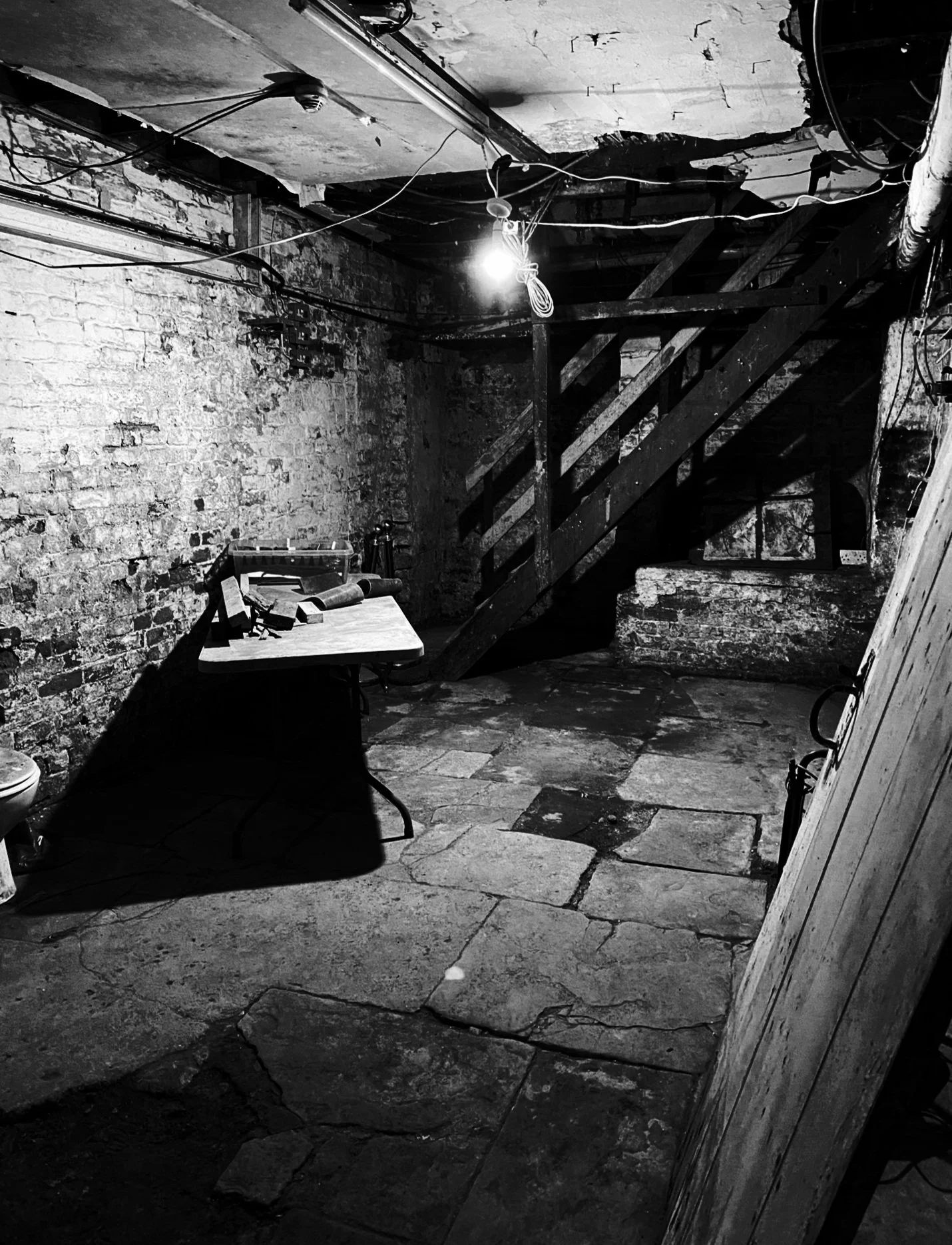 Black and white photo of a basement with exposed brick walls, a wooden staircase, a table with tools, and a single exposed light bulb hanging from the ceiling.