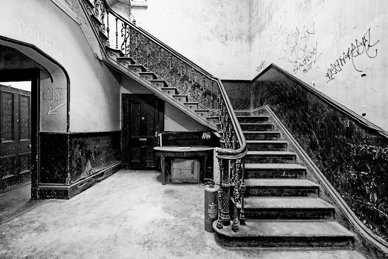 Black and white photo of an old staircase with ornate railing, leading up from a hallway with graffiti-covered walls and a sign indicating men's and women's restrooms.