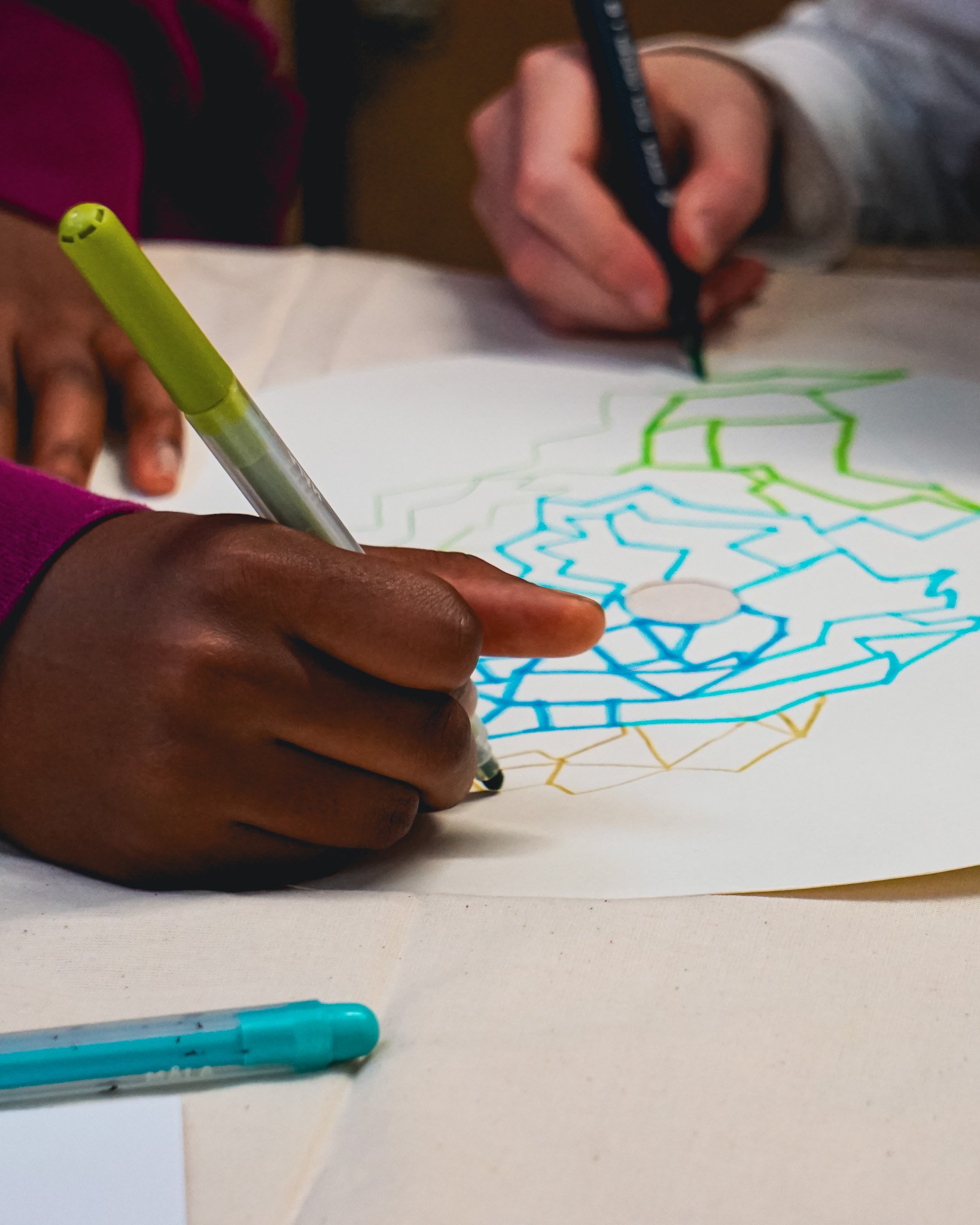 Two people coloring a geometric design on paper with colored pens, focusing on hands and art.