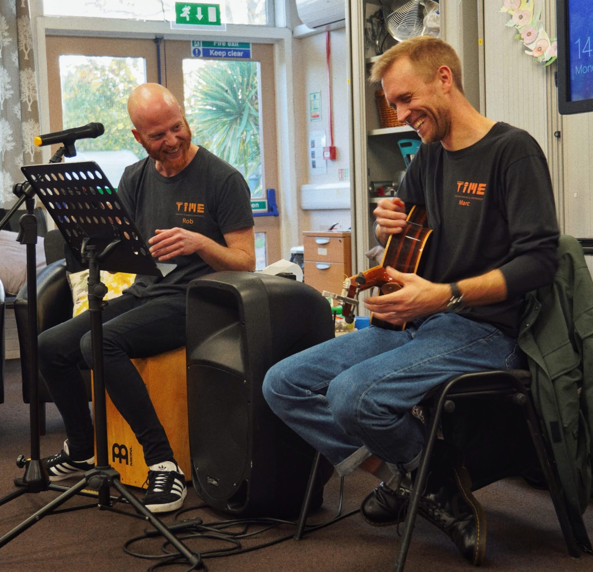 Two men sit at a table, looking at a keyboard with colored stickers, in a room decorated with colorful bowling pin and animal figurines in the background.