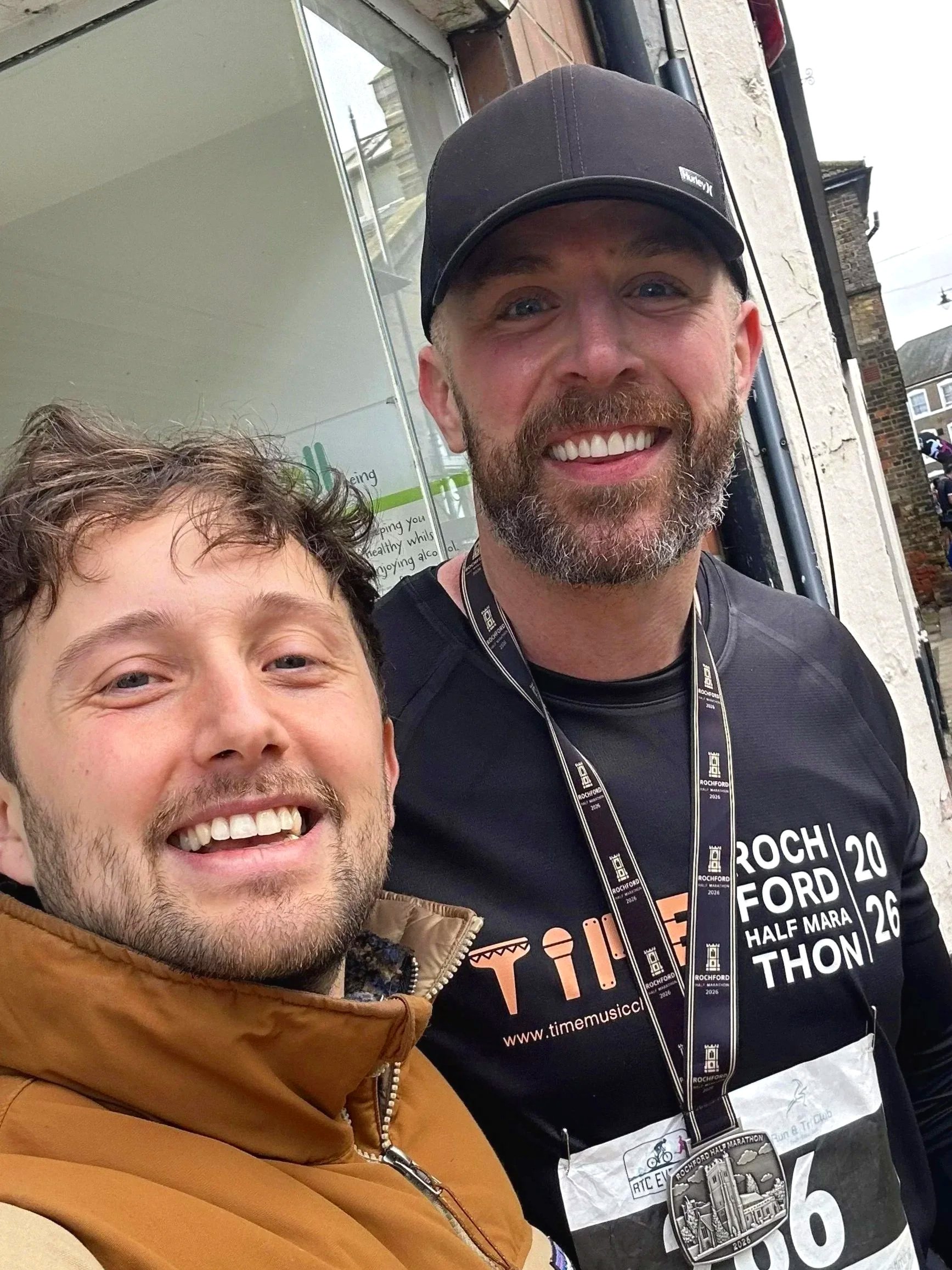Two smiling men taking a selfie outdoors near a building. One man has just completed a half marathon and wears his medal. The other man is from the music charity (TIME) and is celebrating with him.