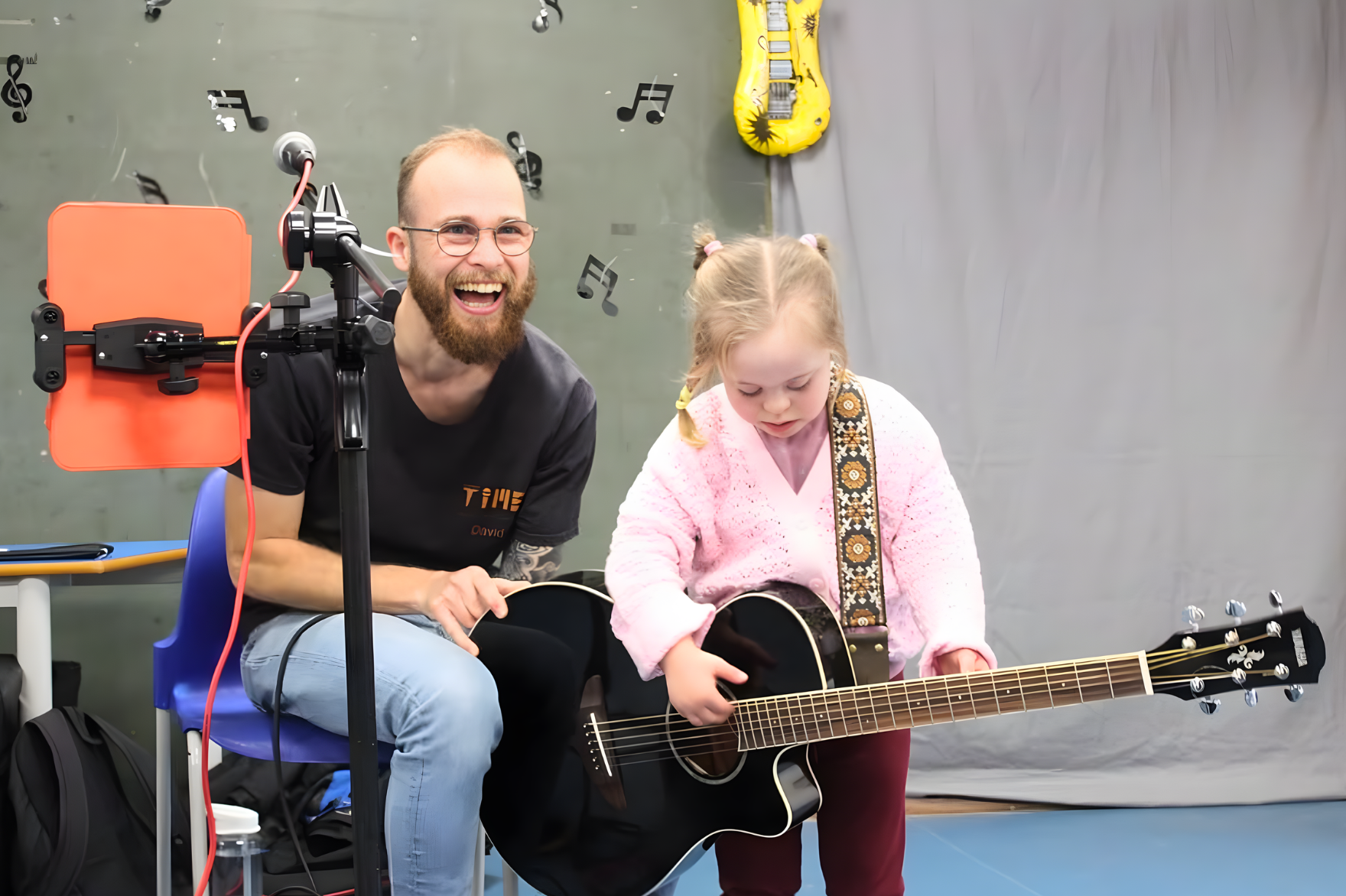 A young girl with blonde hair in pigtails wearing a pink sweater, learning to play guitar with the help of a smiling man with glasses, a beard, and a black T-shirt in a music classroom.