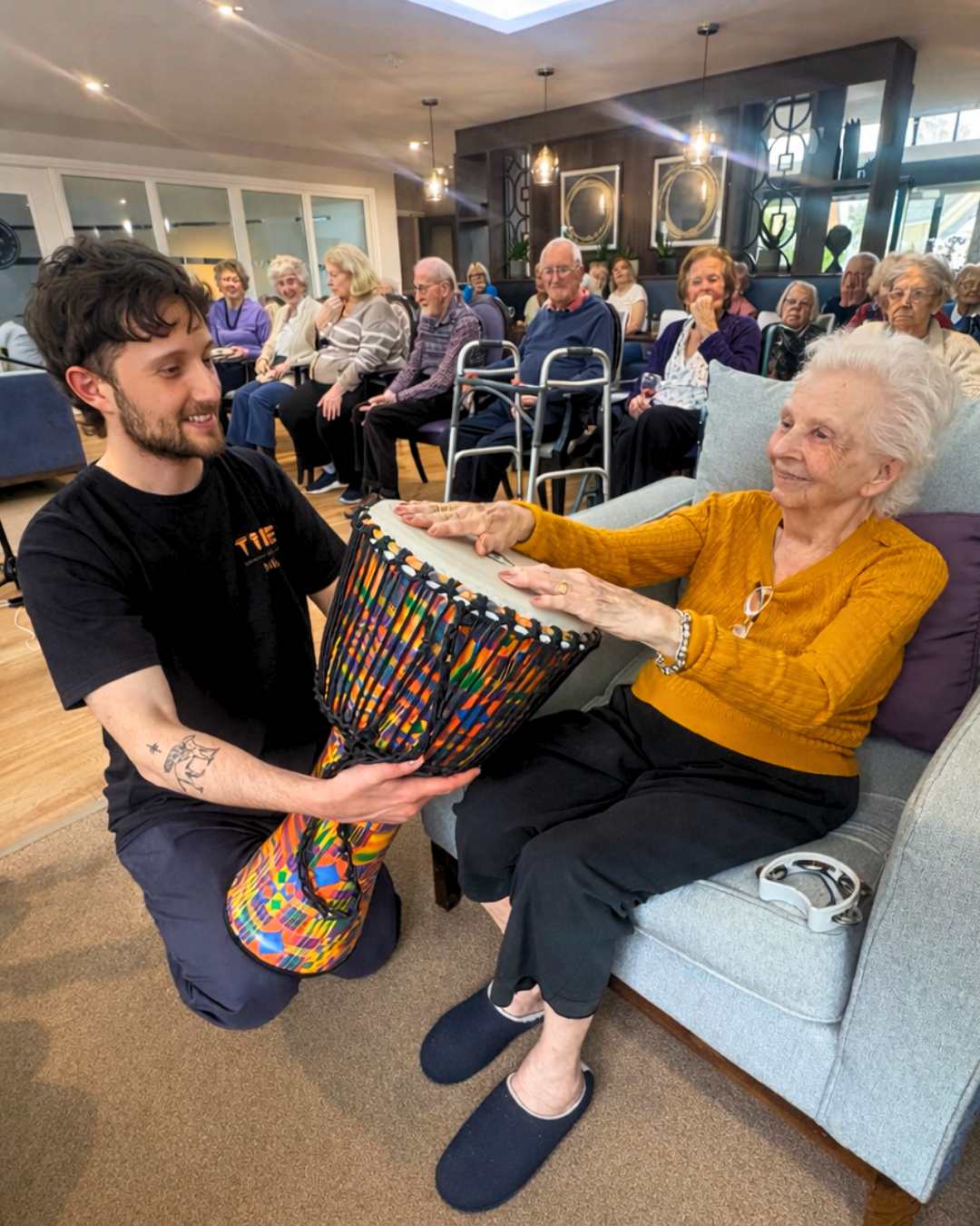 A young man with tattoos on his arm is kneeling and giving a brightly colored djembe drum to an elderly woman sitting on a sofa. The elderly woman is reaching out to accept the drum, smiling, in a room with several other elderly people sitting and watching, some with walkers, in a well-lit, cozy setting.