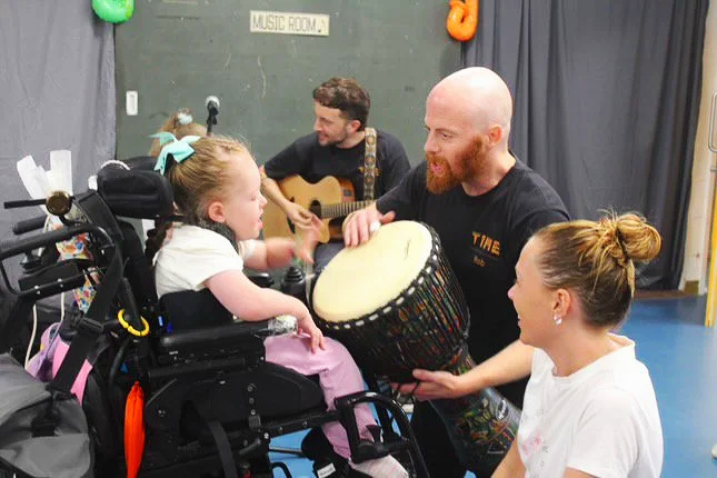 A group of four people, including a young girl in a wheelchair, a woman, a man with a guitar, and a man holding a drum, in a music room, engaging in music activities.