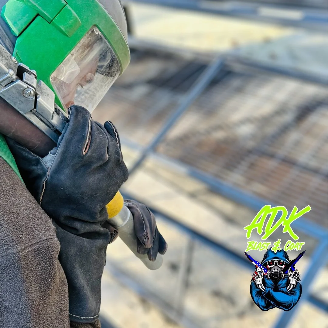 A person wearing a green helmet, black gloves, and a brown jacket holding a spray gun, with an outdoor construction or roofing site in the background. There is a logo in the bottom right corner that says "AUX Blast & Coat" with an illustration of a masked figure wearing sunglasses and a hoodie, holding spray guns in each hand.