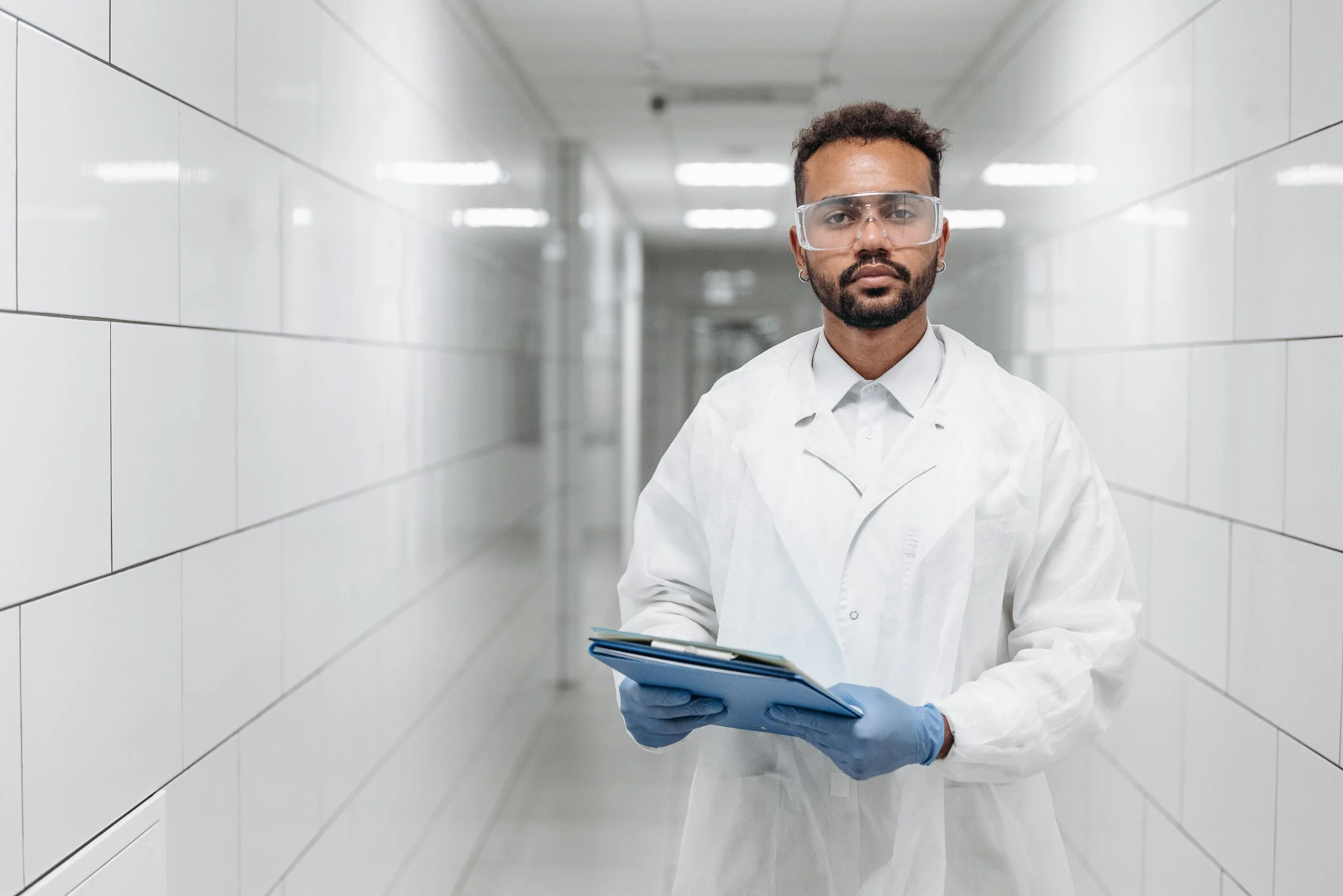 A researcher in a white coat and gloves walking a clinical corridor with a tablet