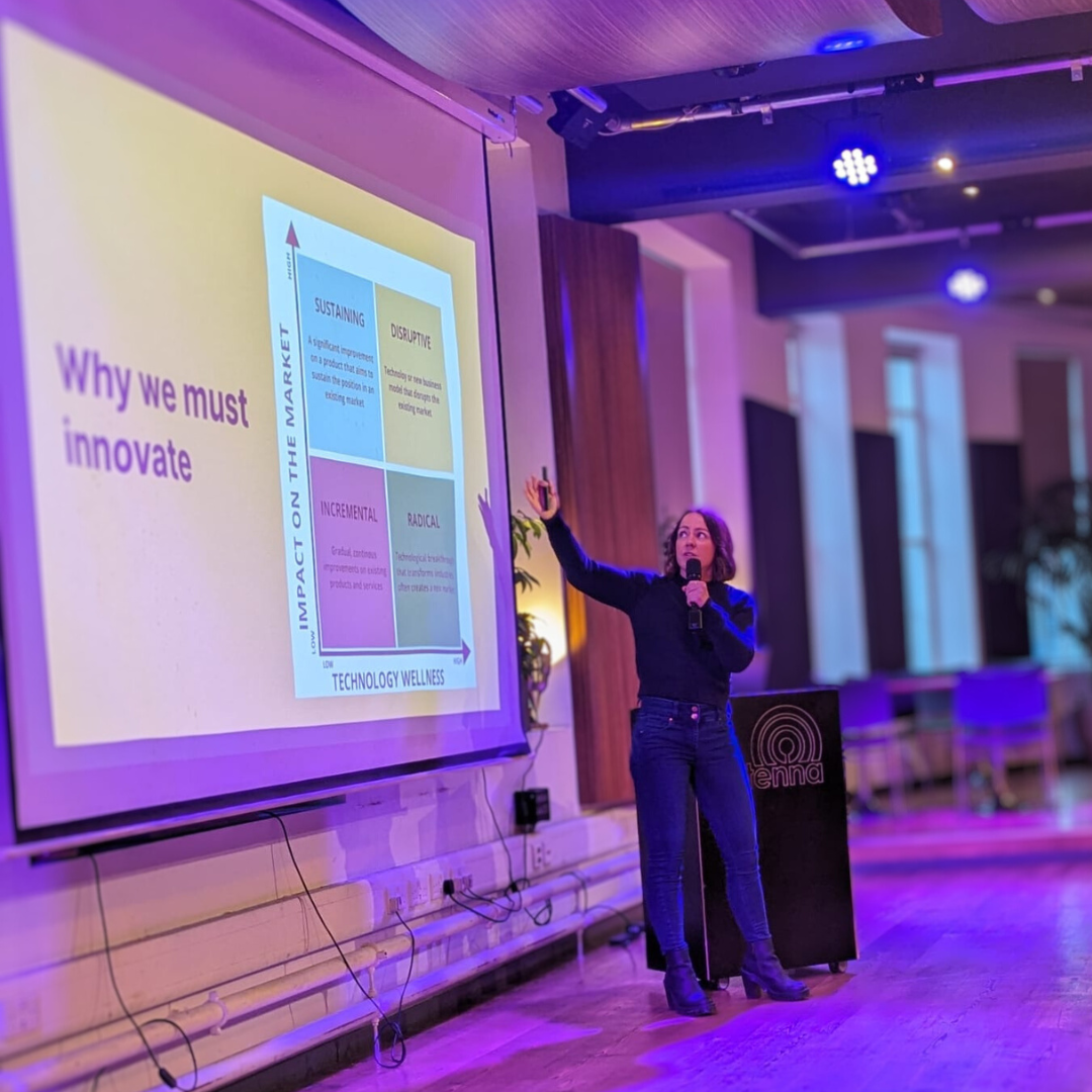 Dr Chloe Sharp presenting to an audience, pointing to a slide on why we must innovate