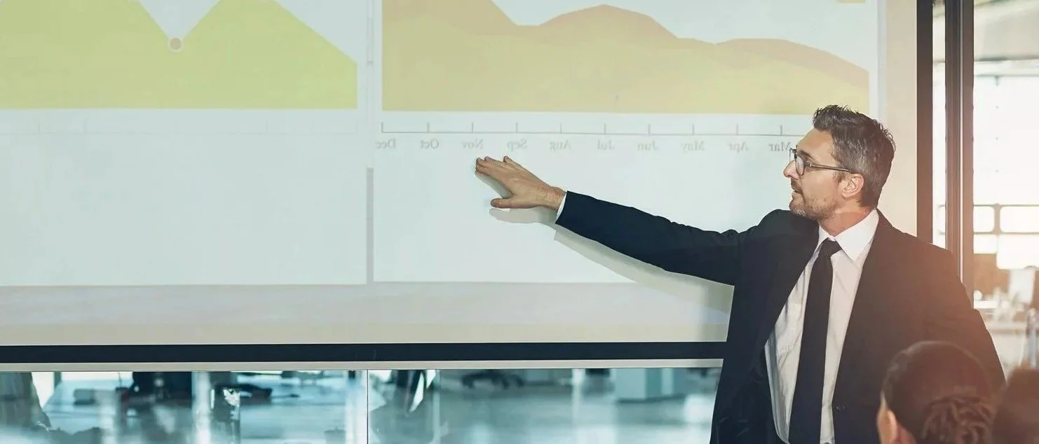 A presenter pointing to financial charts on a screen in a boardroom