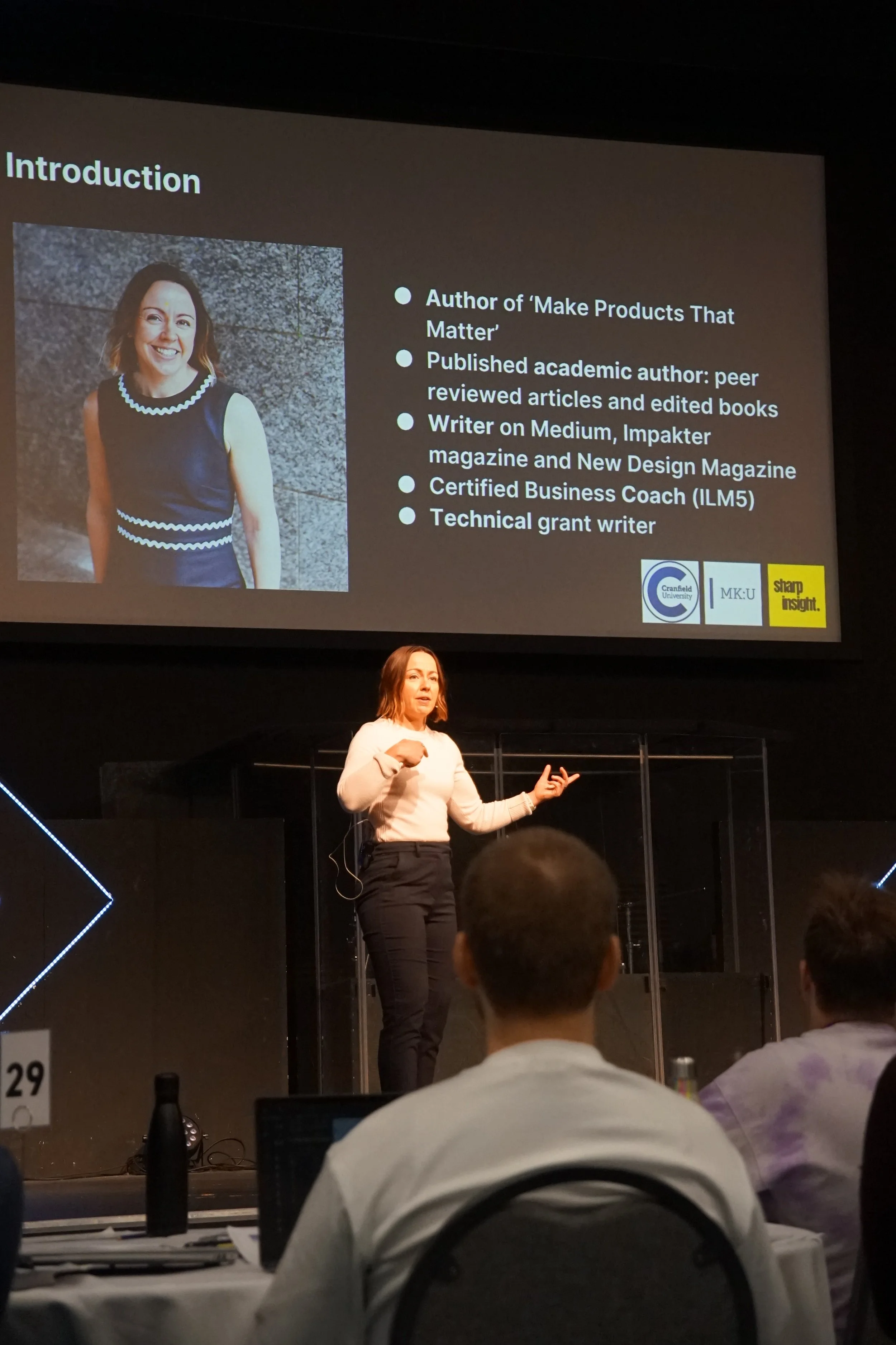 A woman giving a presentation on stage in front of a large screen displaying her biography and credentials, while seated audience members watch.