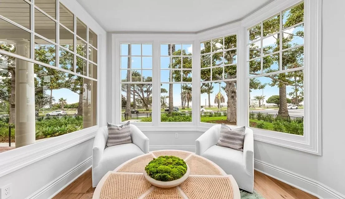 Sunlit room with large windows, two white armchairs, a round coffee table, and a view of trees and blue sky outside.