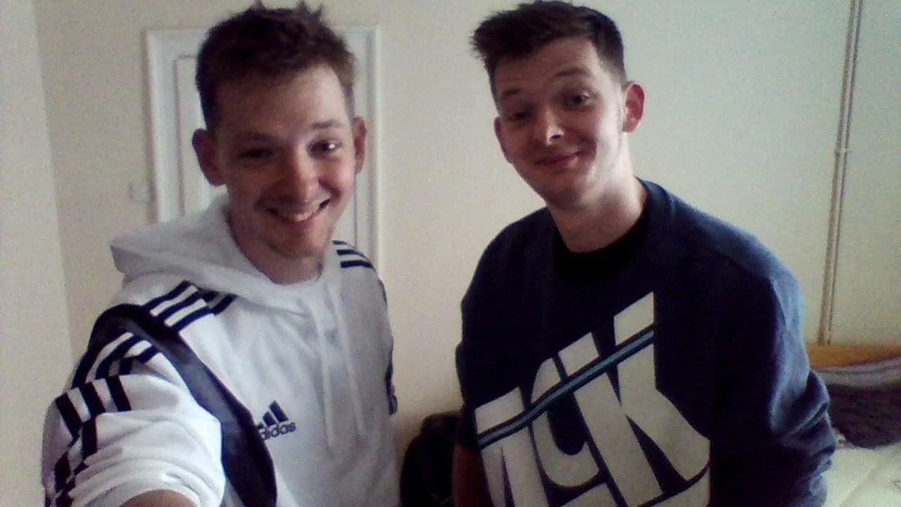 Two young men taking a selfie indoors, smiling at the camera. One wears a white Adidas hoodie, and the other wears a black Nike sweatshirt.