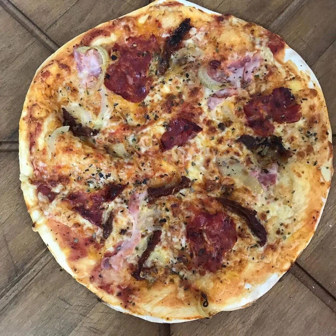 A whole pizza topped with cheese, pepperoni, ham, onions, and herbs on a wooden surface.