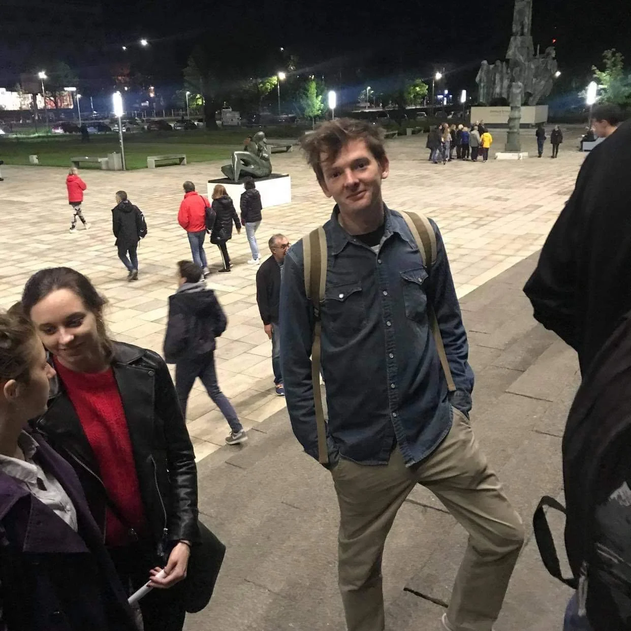 A group of young people standing and talking in a city square at night, with a large sculpture and trees in the background.