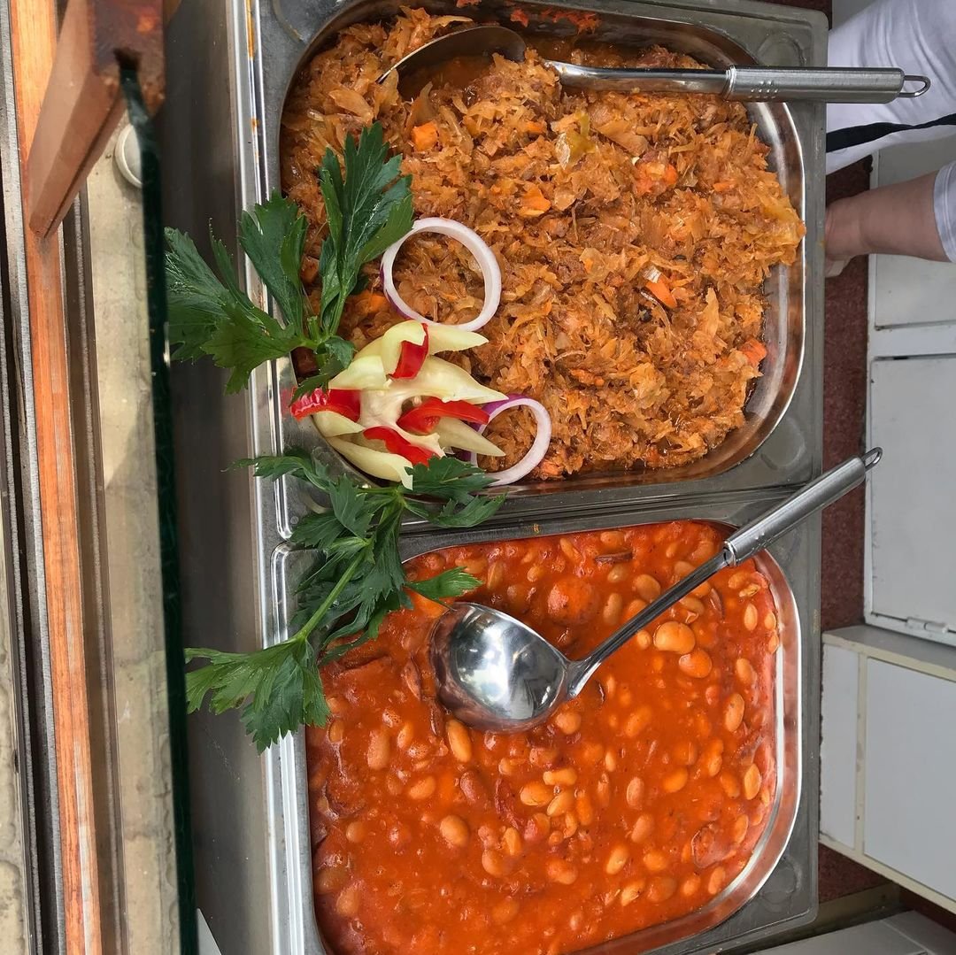 Tray of cooked food with two sections: the top section contains shredded meat with vegetables, garnished with parsley, sliced onions, and sliced red and yellow peppers; the bottom section contains beans in a tomato sauce. There are serving spoons in 